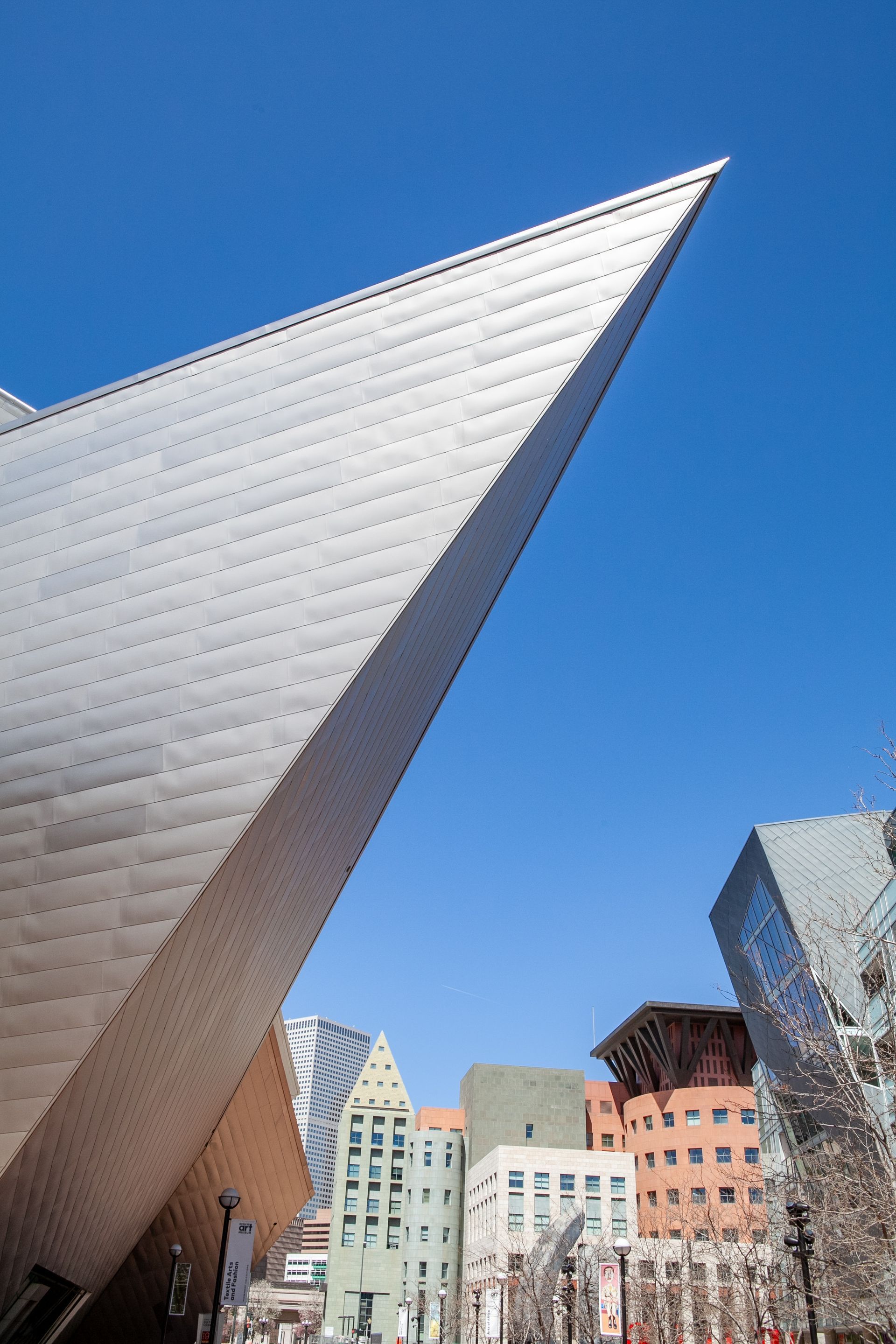 Angled silver modern sculpture against blue sky with colorful buildings below