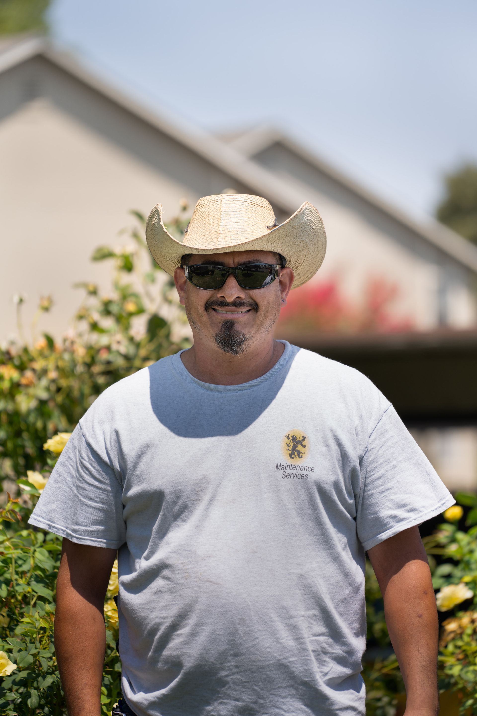 Man wearing a straw cowboy hat and sunglasses, standing in front of a house and bushes.
