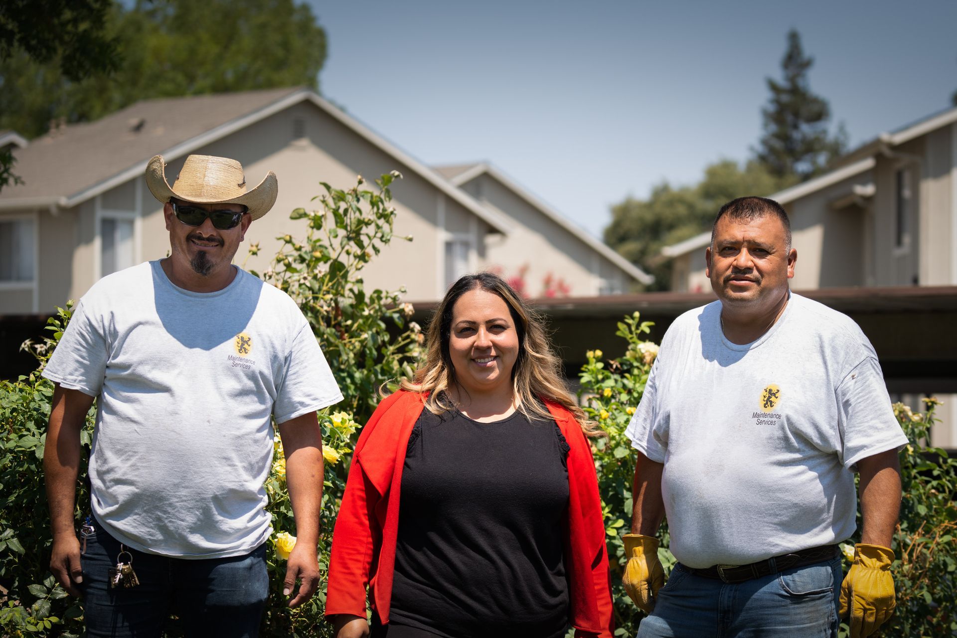 Three people smiling outdoors in front of bushes and apartment buildings. Two men wear white t-shirts; one has a hat.