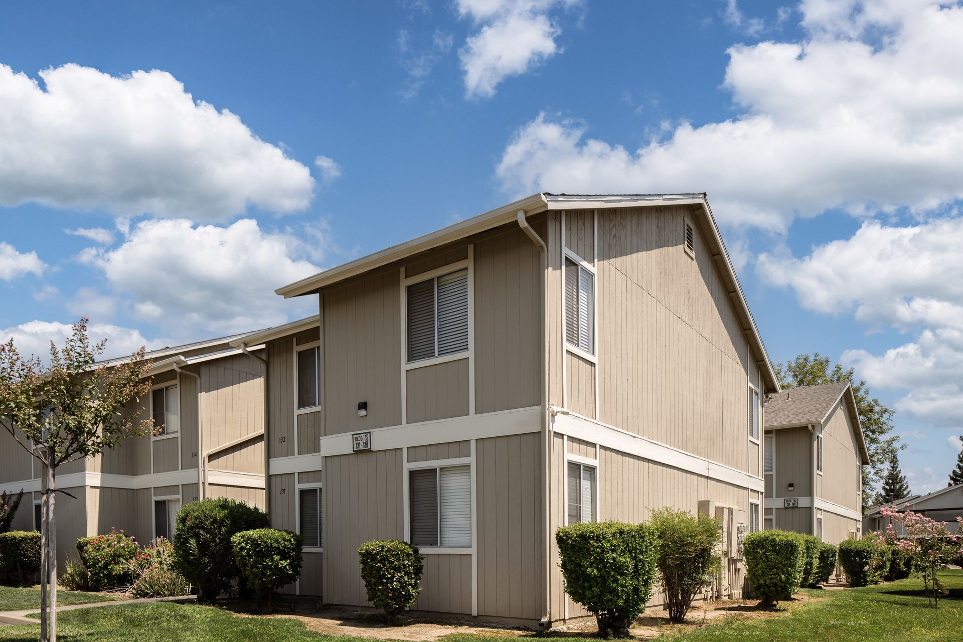 Two-story beige apartment buildings with green bushes, blue sky, and white clouds.