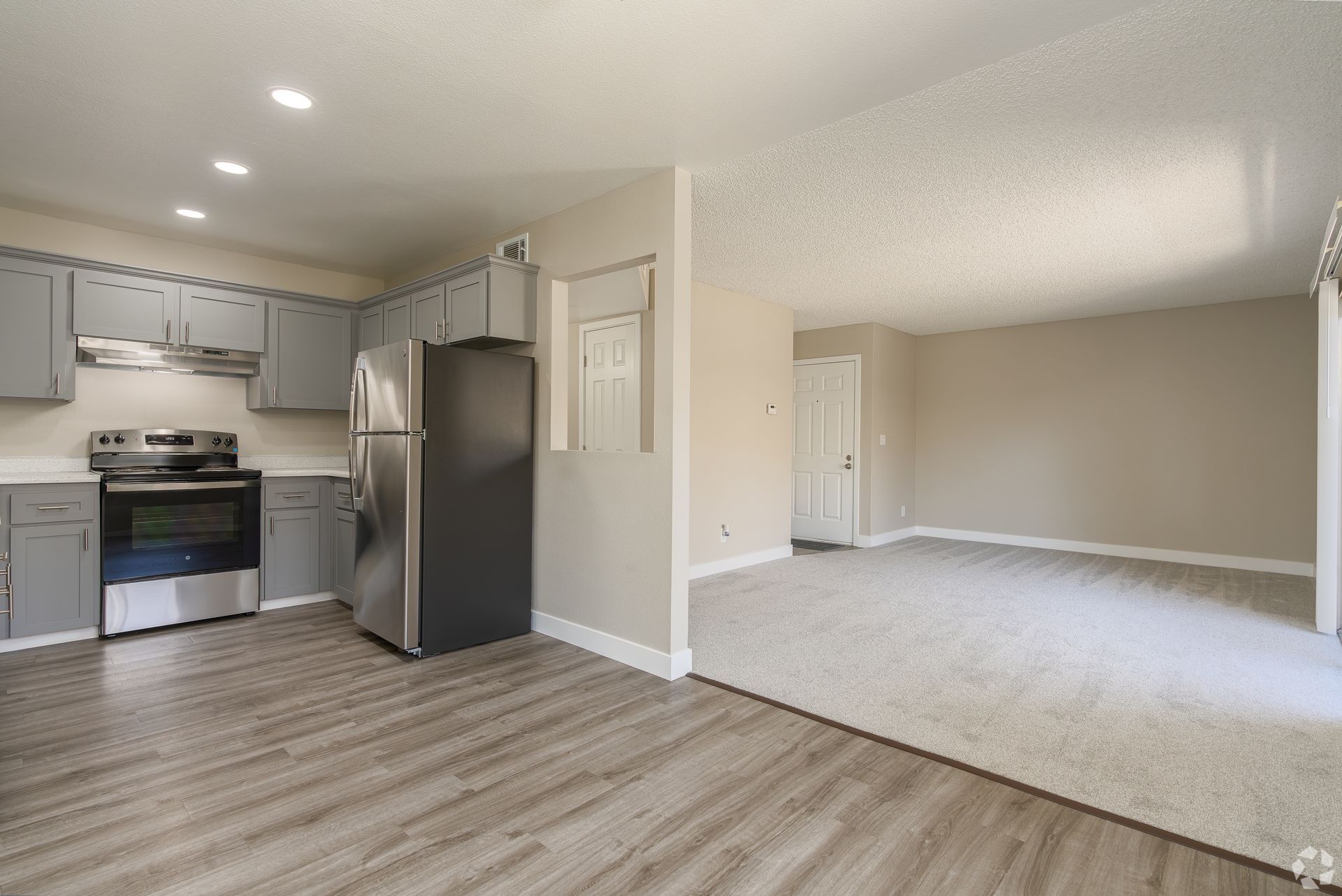 Kitchen and living room in apartment with gray cabinets and appliances, beige walls, and light gray carpet.