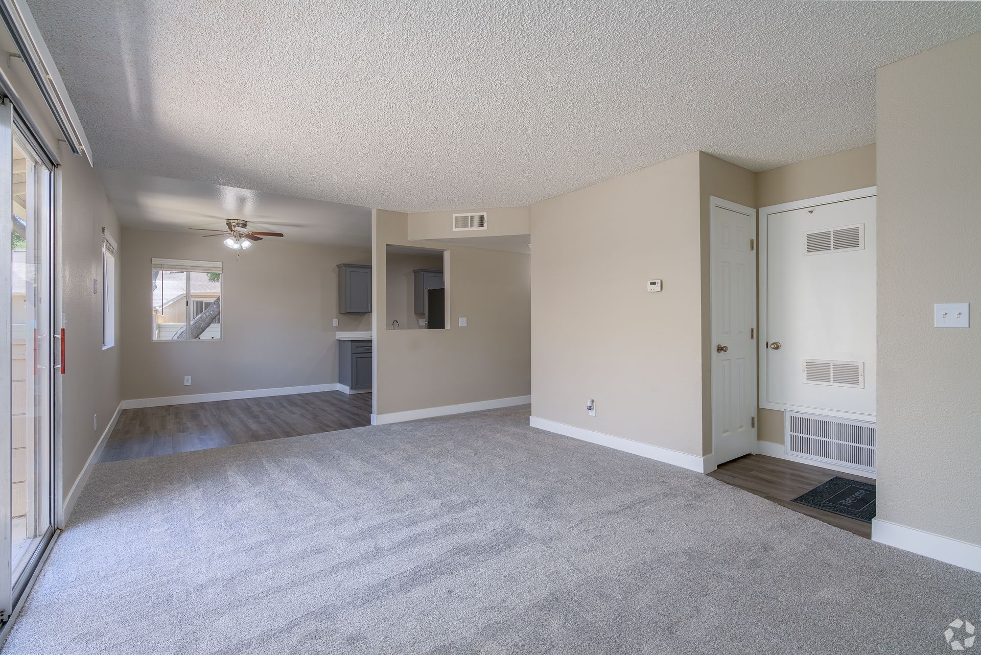 Empty living room with gray carpet, neutral walls, and a ceiling fan. Door on the right.