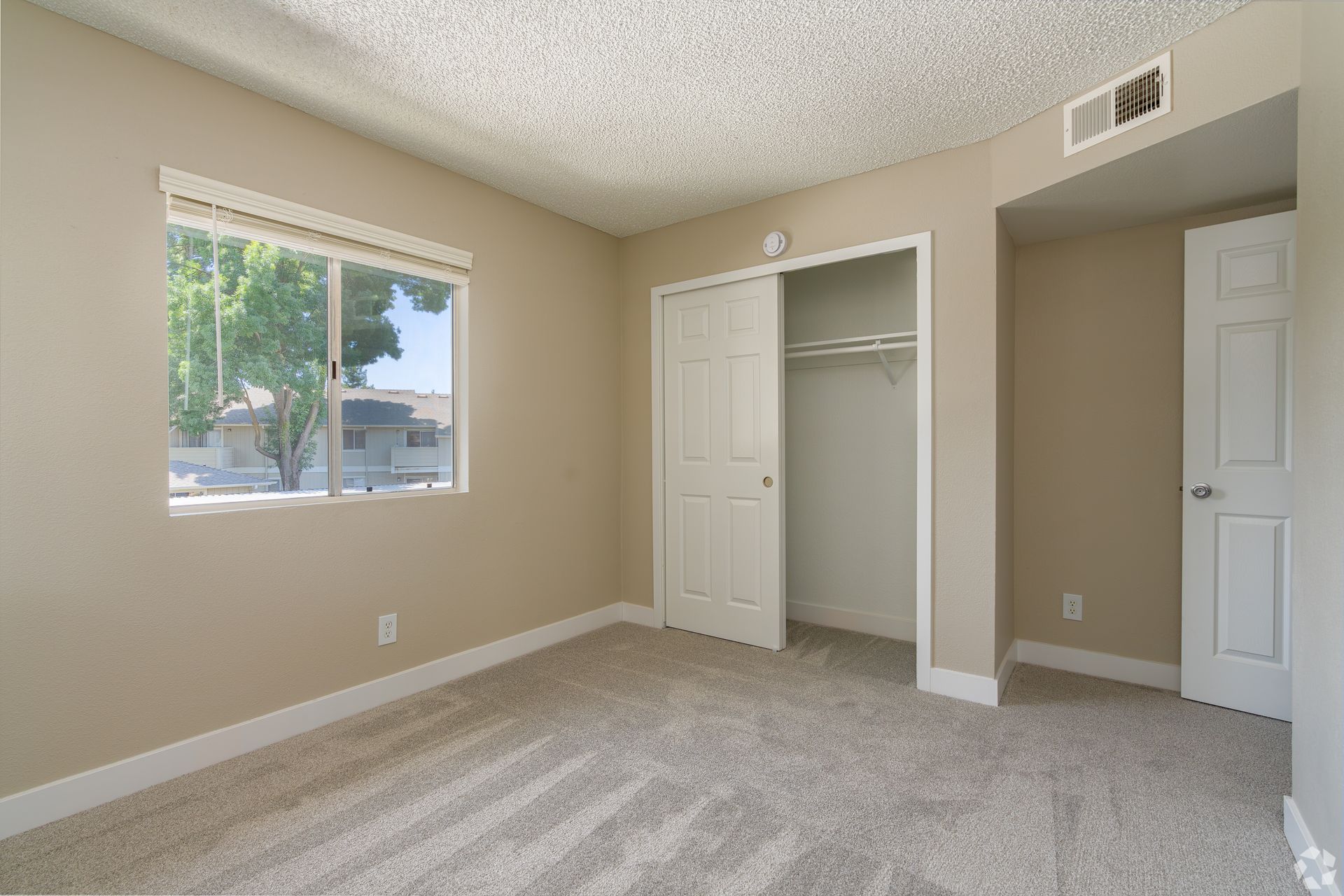 Empty beige bedroom with a window, closet, and door; light gray carpet.