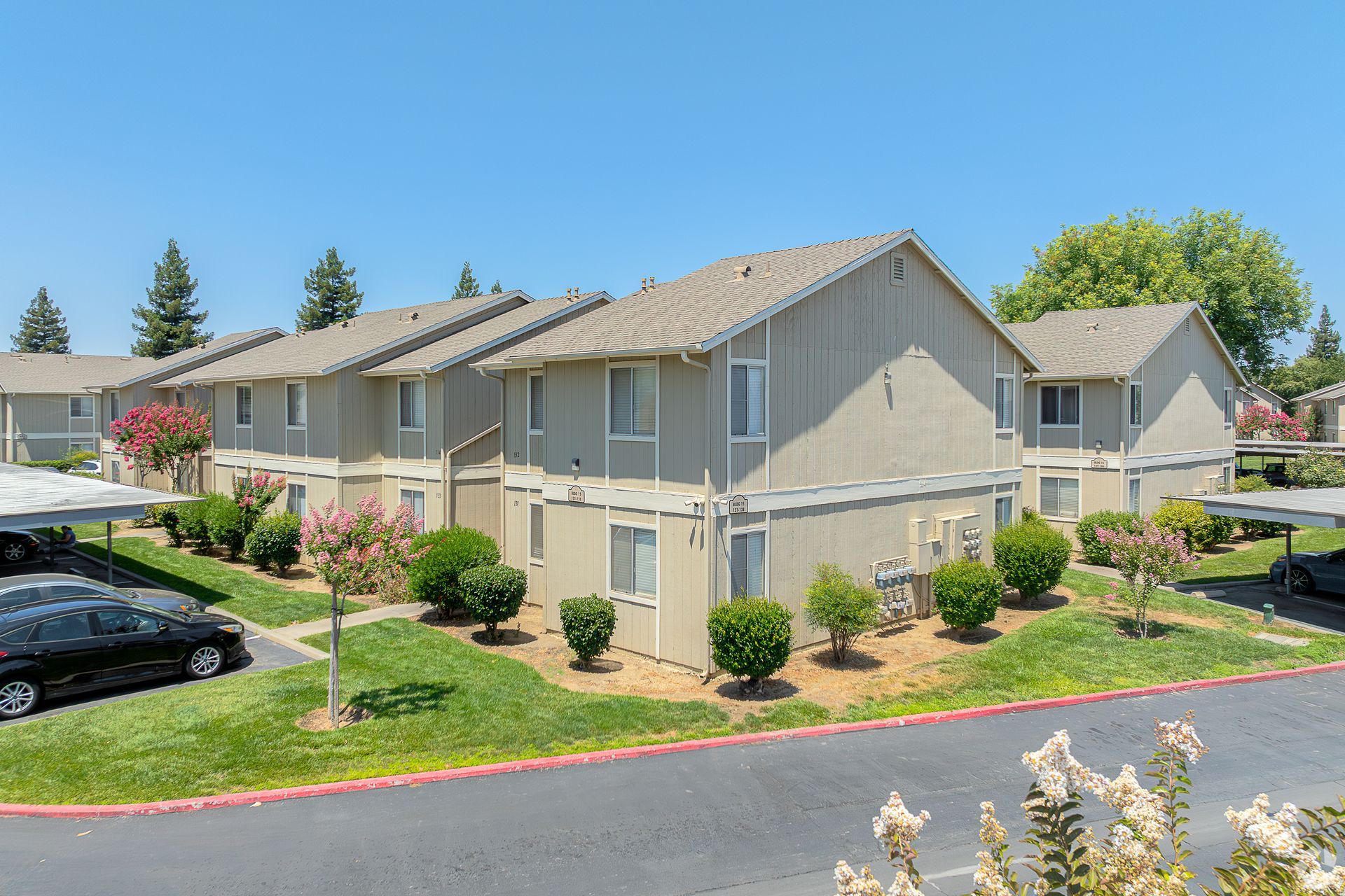Apartment complex with tan buildings, green lawns, blue sky, and parked cars.
