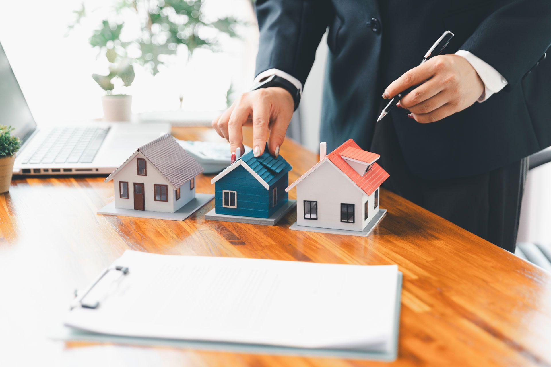 Person in suit points to miniature blue house on desk with other model houses and paperwork.