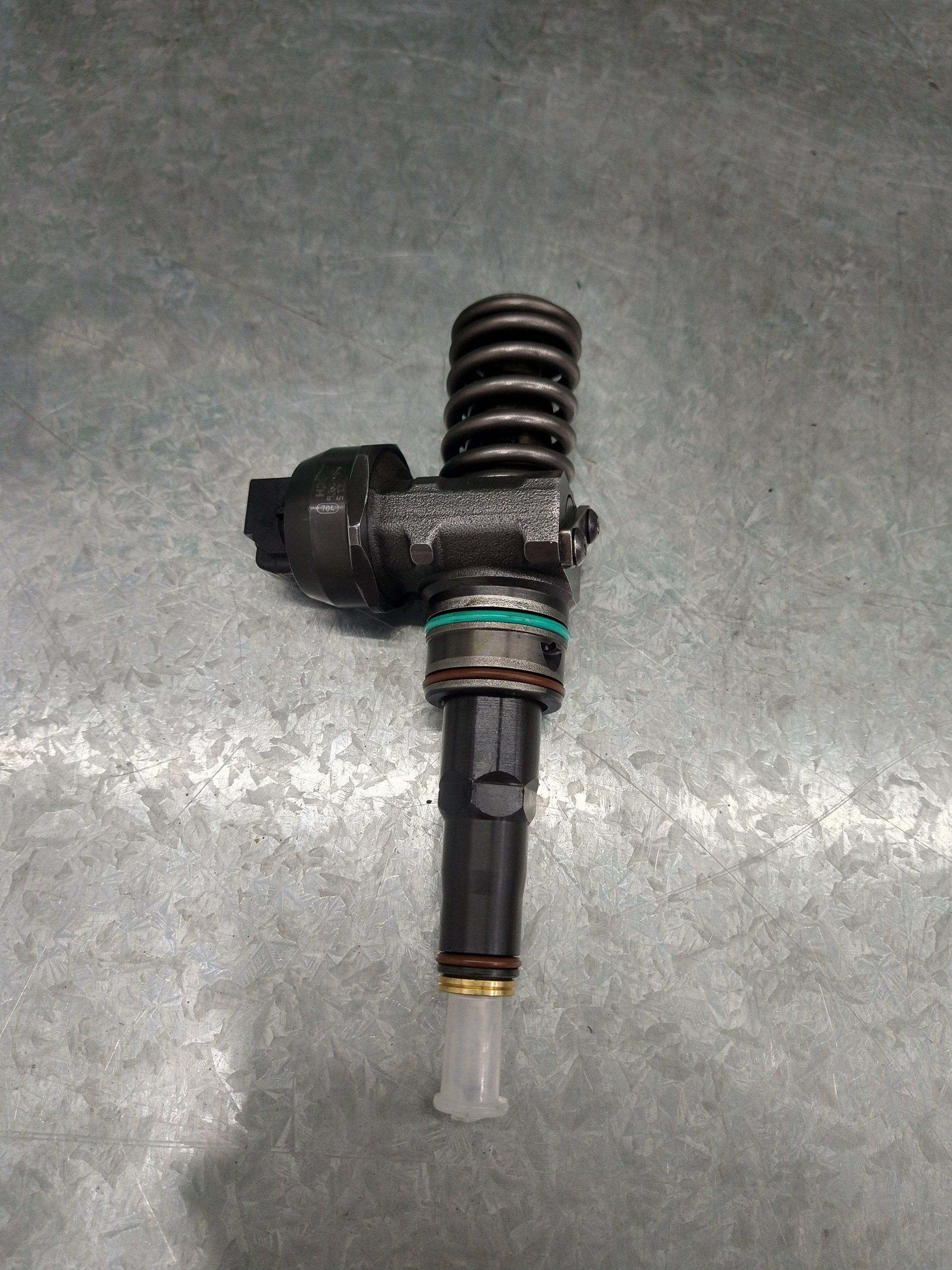 Inyector de combustible con cuerpo negro, resorte y punta transparente, sobre superficie metálica.