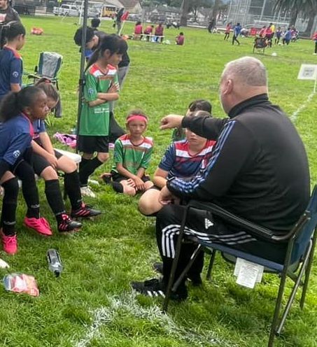 A coach sitting in a chair talking to a group of children on a soccer field.