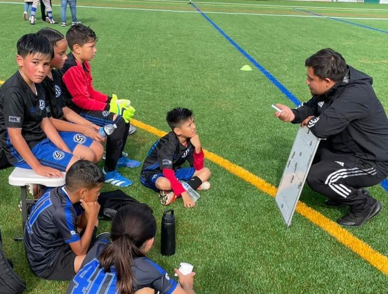 A group of young boys are sitting on a bench on a soccer field  while being coached,