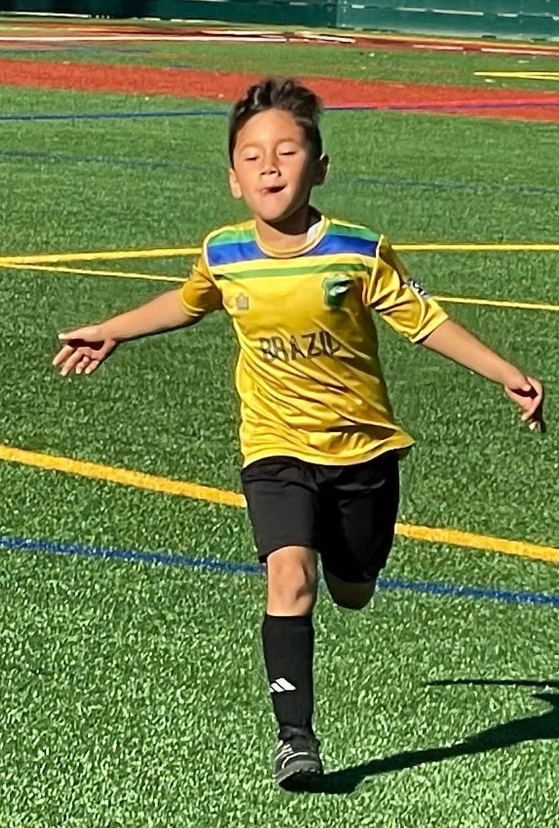 A young boy is running on a soccer field with his arms outstretched.