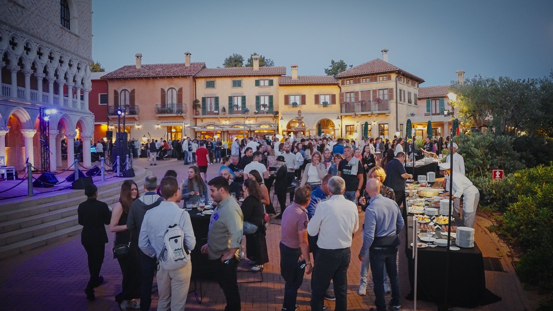 Crowd at an outdoor evening event in a European-style square; people mingle, some near a food station.