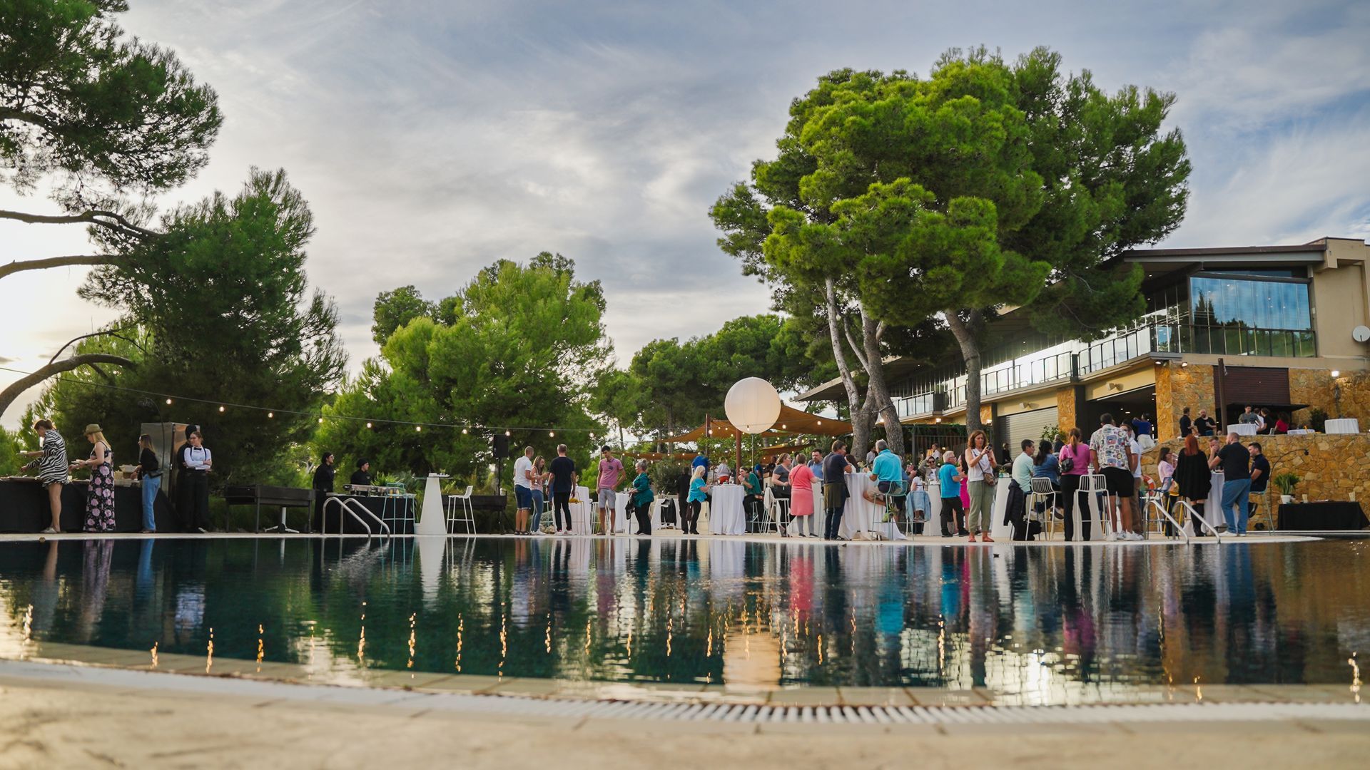 Wedding reception by a pool. Guests gather, a large balloon floats, reflecting in the water. Sunny, outdoor setting.