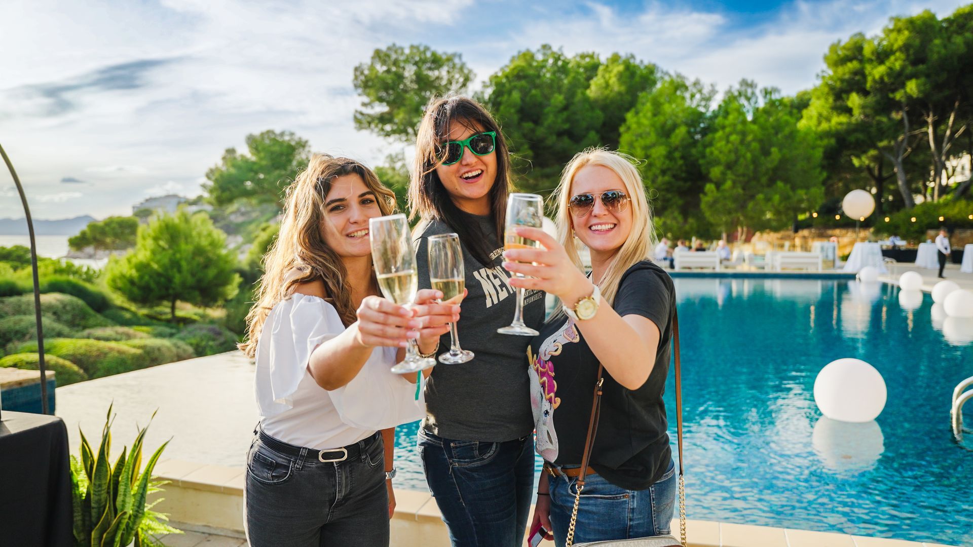 Three women toasting champagne by a pool. Bright sunlight, smiling faces, outdoor event.
