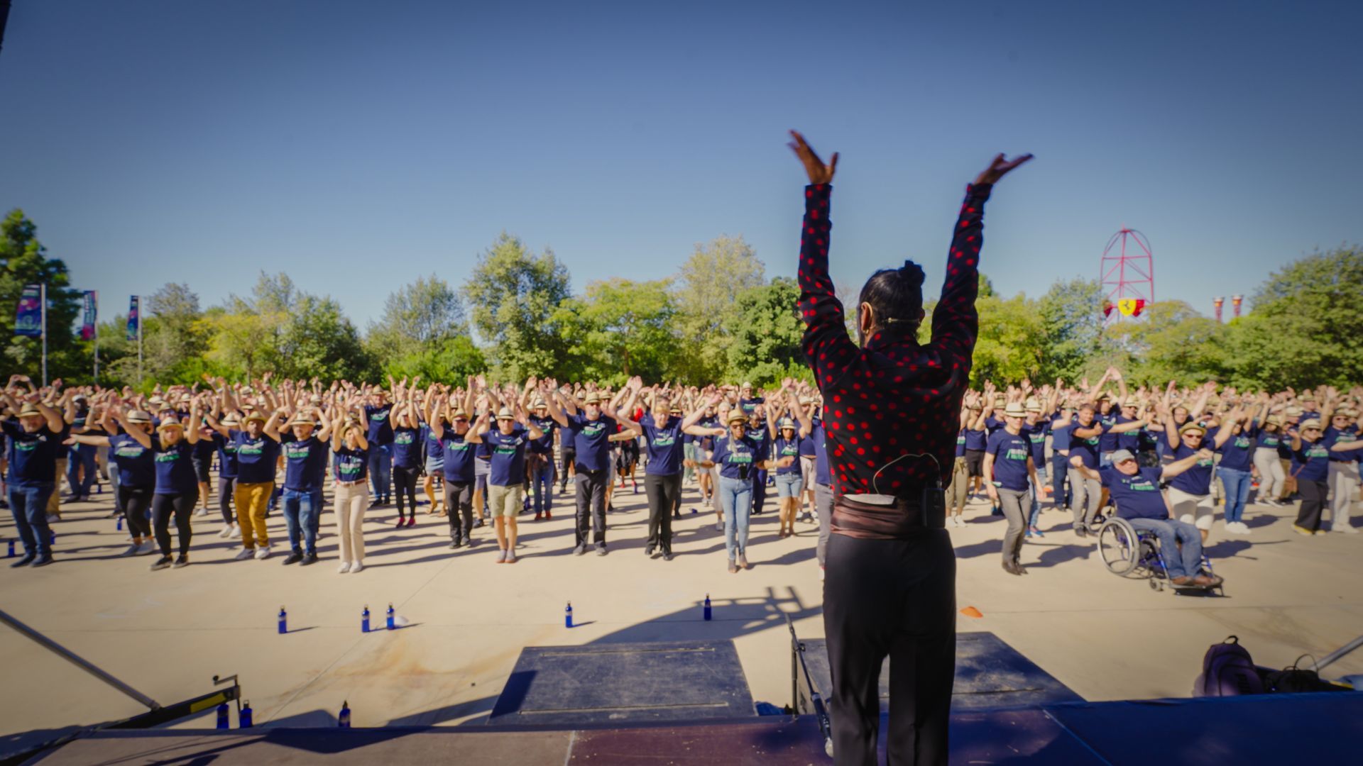 Woman on stage leading group in outdoor exercise; arms raised. Large crowd mirroring pose.