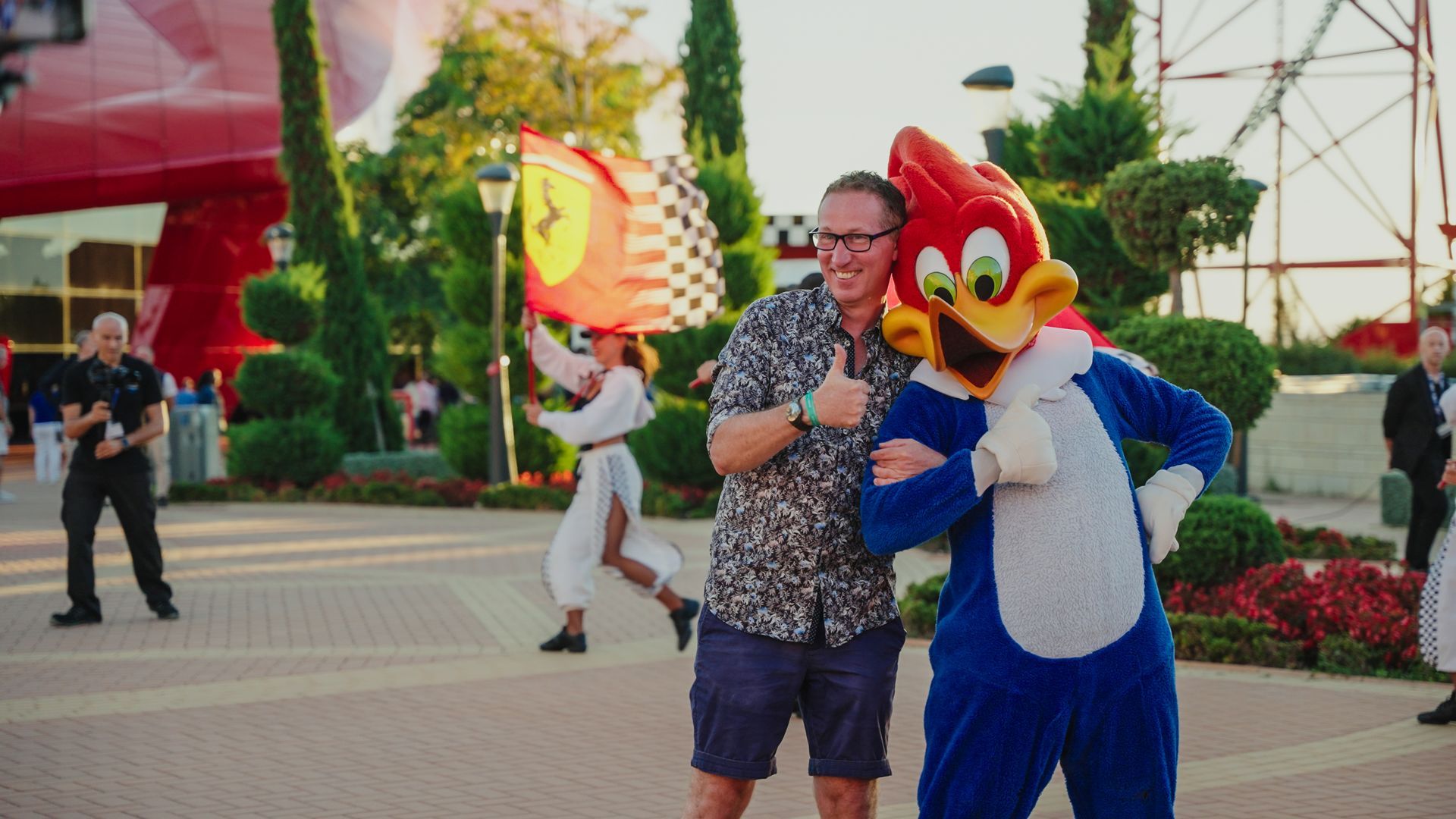 Man with Woody Woodpecker character, thumbs up. Red Ferrari flag in background, outdoor setting.