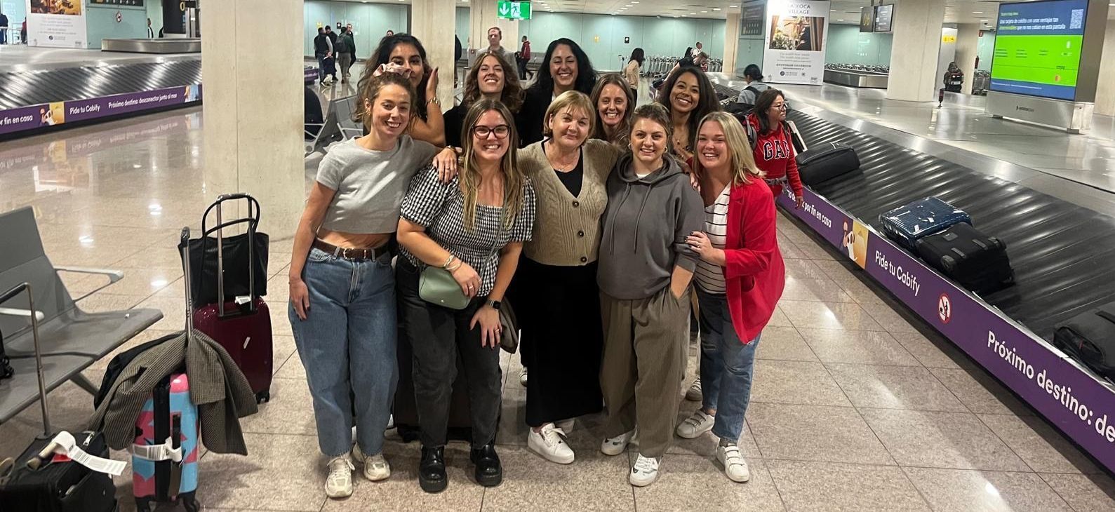 Group of women at an airport baggage claim, smiling, some with arms raised.