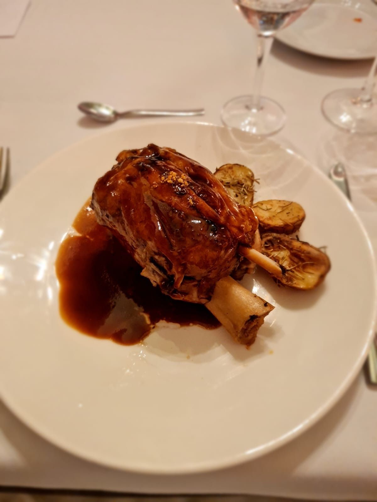 Braised lamb shank with sauce and roasted potatoes on a white plate, formal dining setting.