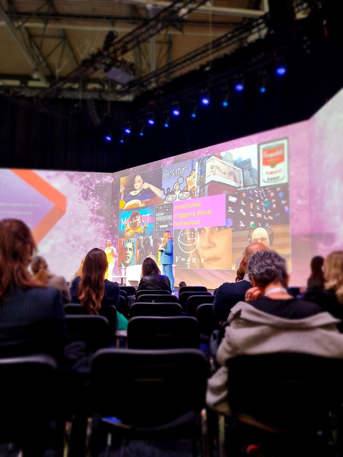 Audience watches a presenter on stage at a conference. Large screen displays images. Rows of chairs.