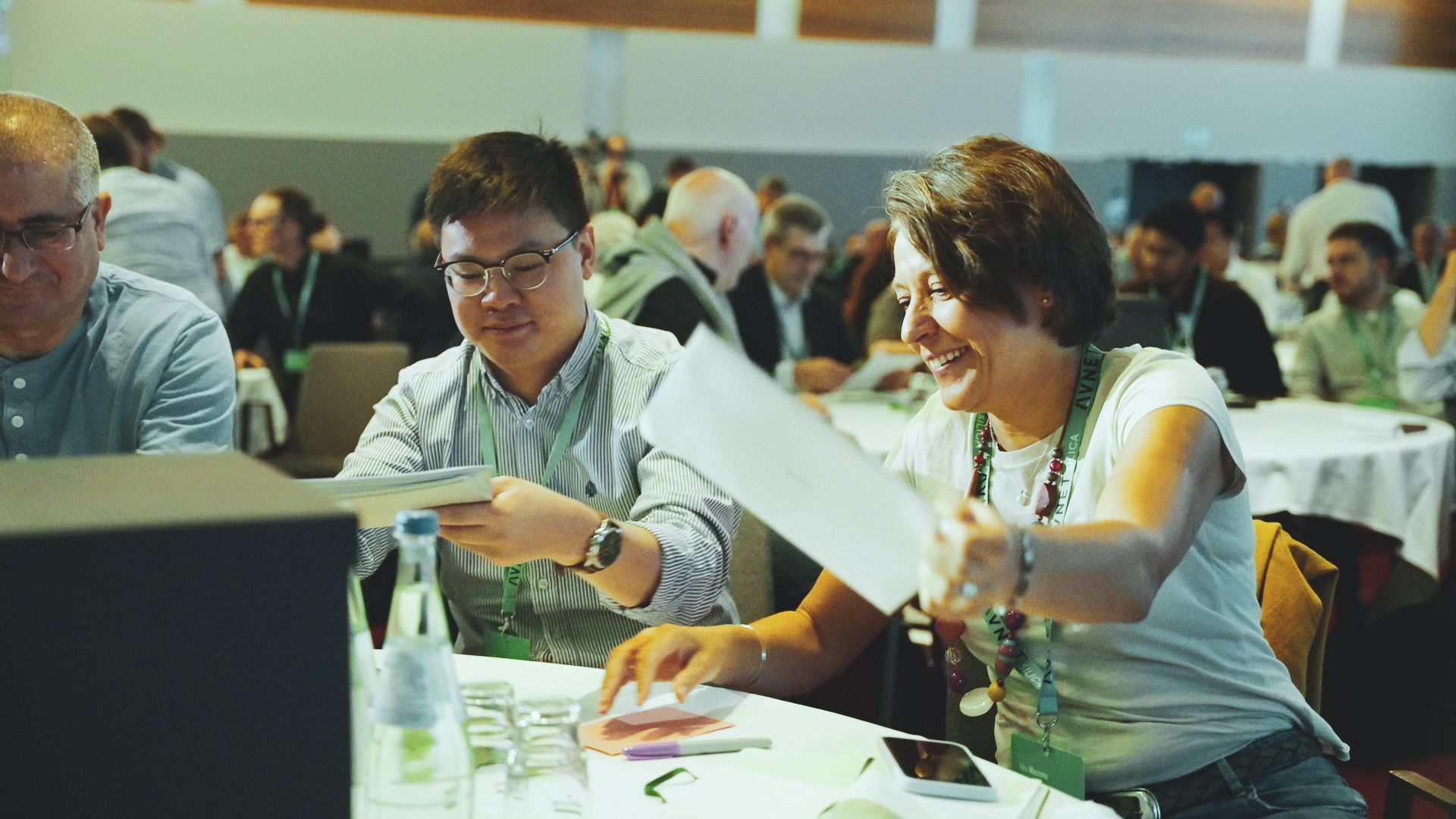 Two people at a conference table, sharing papers. Woman smiles as she hands the man a paper.