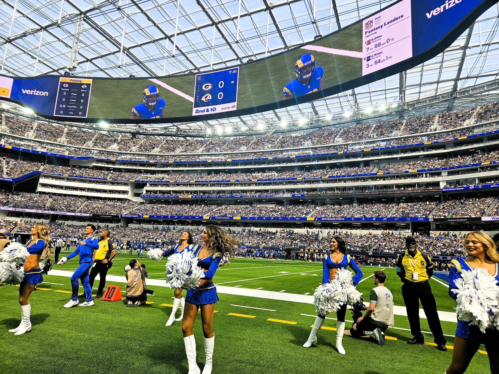 Cheerleaders on a football field in a packed stadium. They wear blue and white. Fans watch in the background.