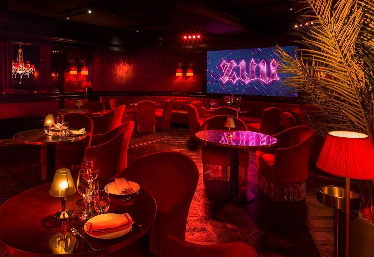 Red-lit, elegant restaurant interior. Tables set with dishes, chairs, and a large screen displaying the word “Zouk.”