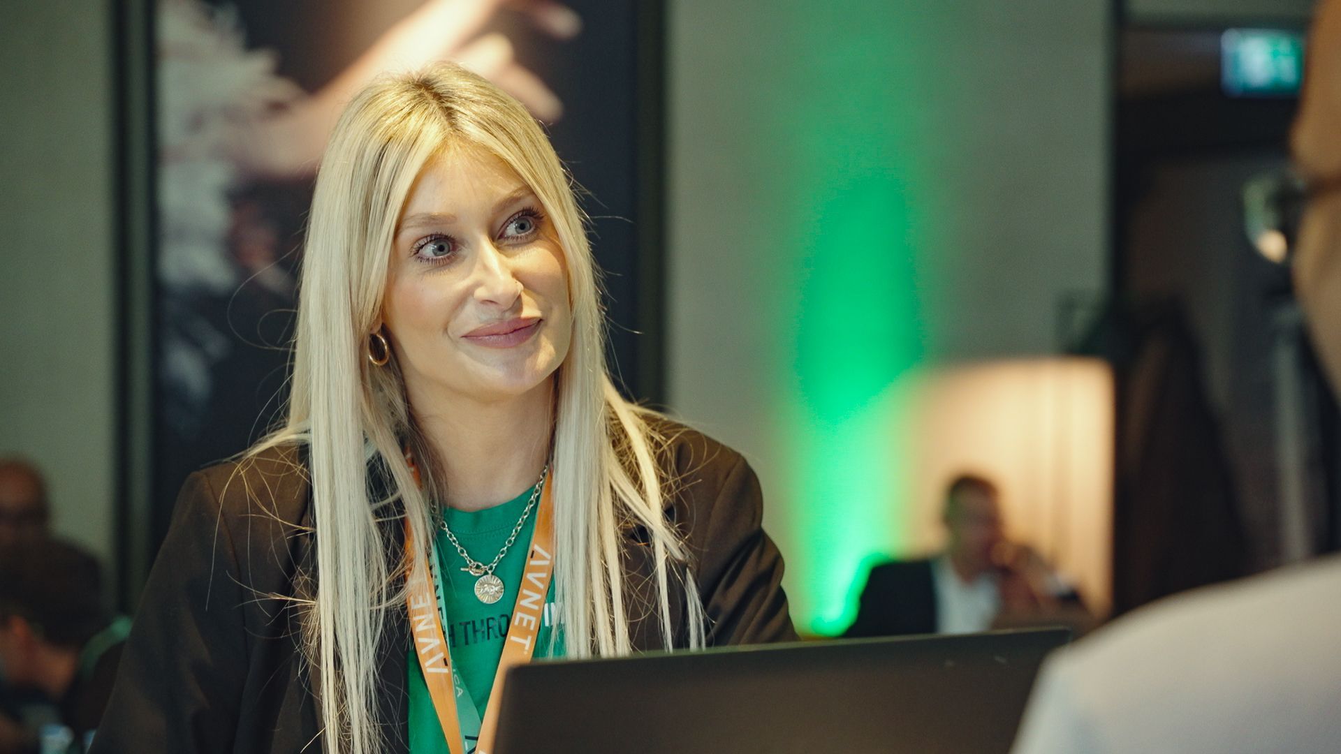 Blonde woman in black blazer smiles, looks towards a person off-screen. She's in a brightly lit setting.
