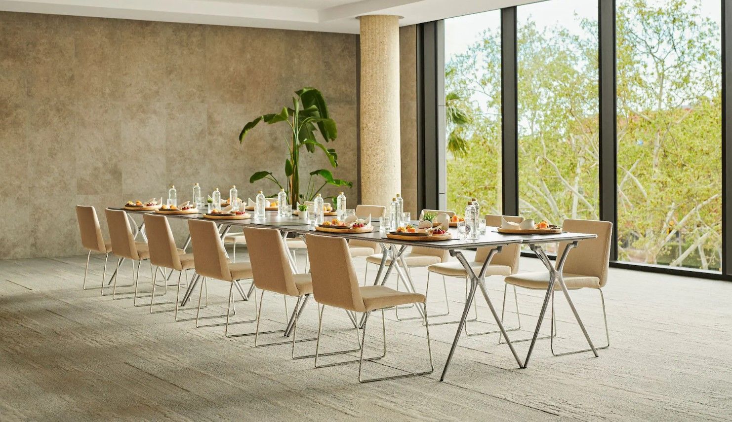 A long table set for a meal with chairs, in a room with large windows overlooking trees.