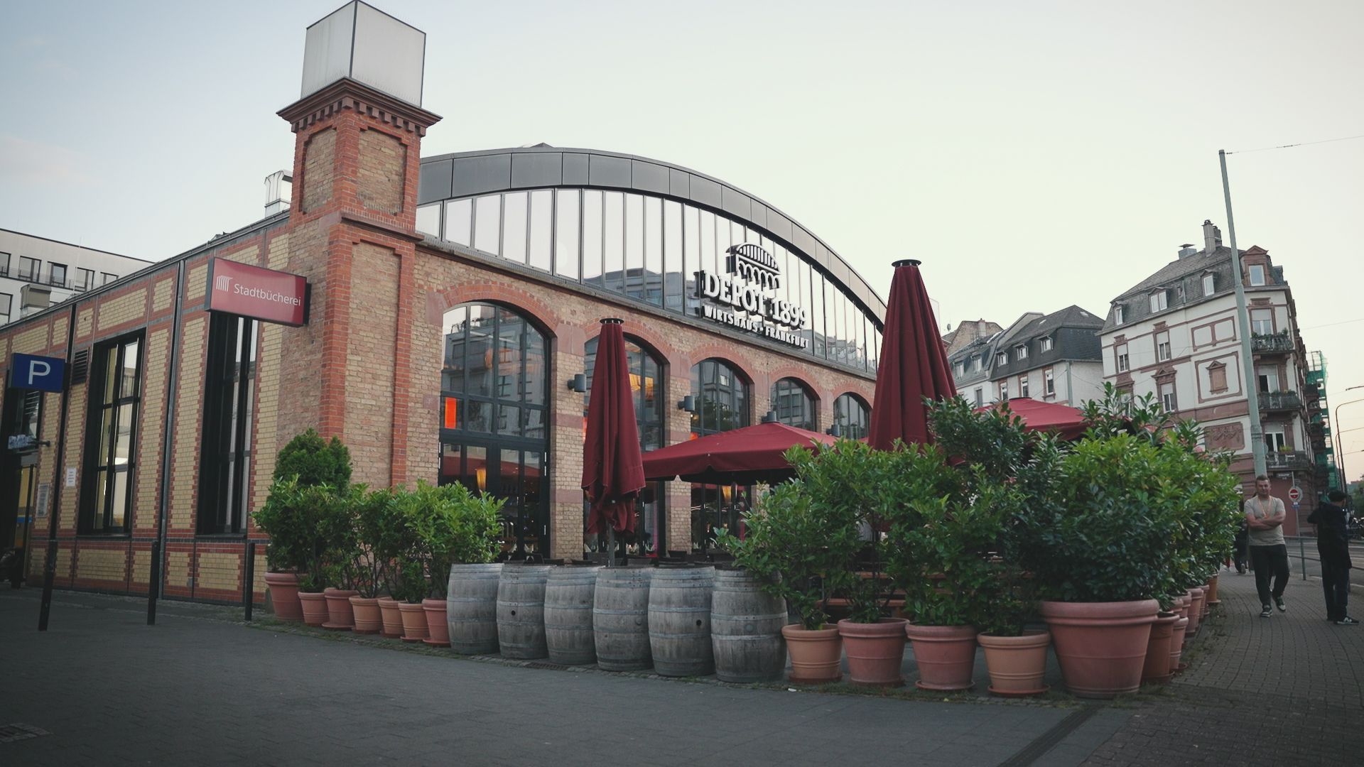 Restaurant exterior with brick facade, large windows, red umbrellas, and potted plants.