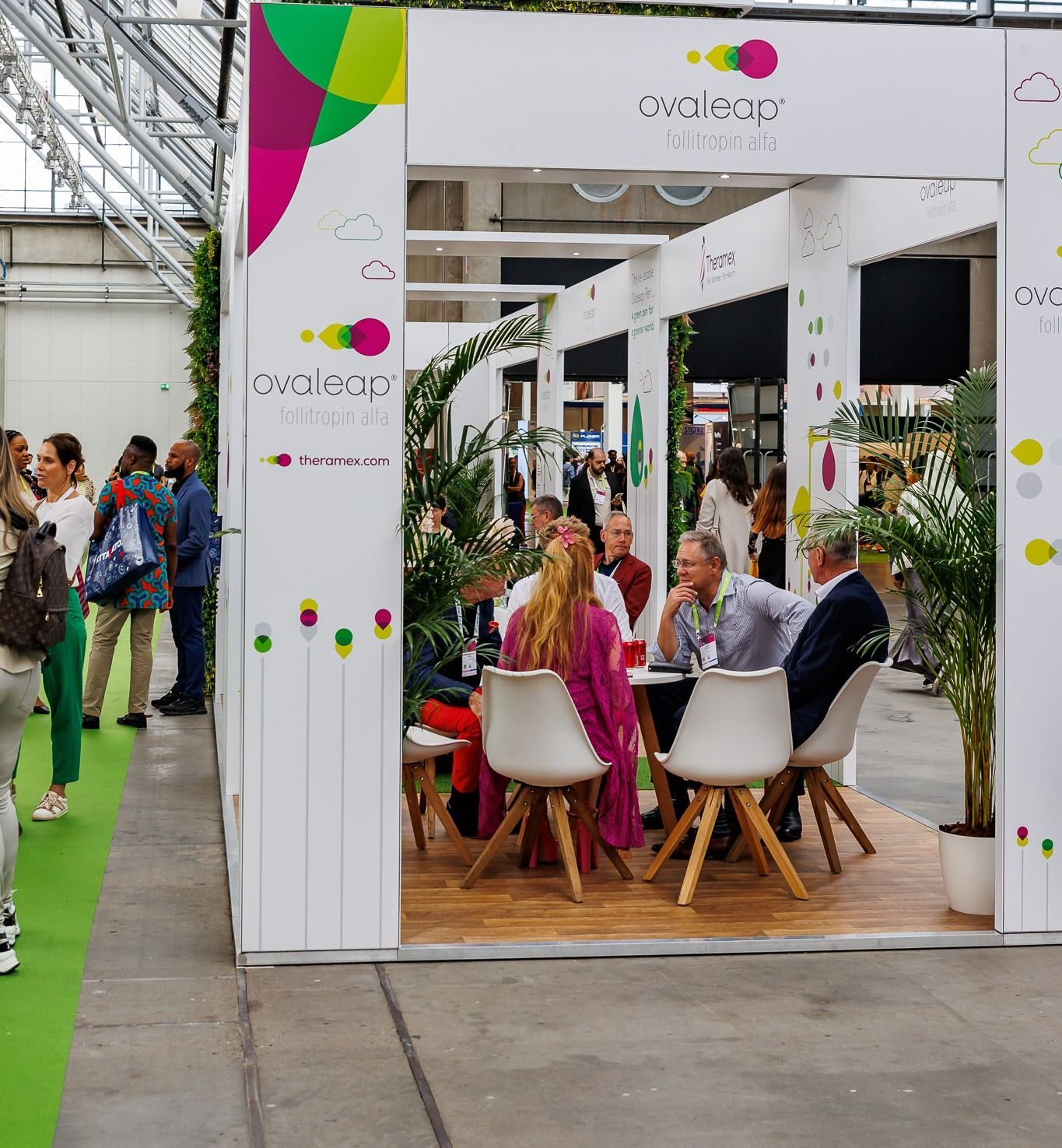 People seated at a table in a booth with branding, plants, and other attendees at a convention.