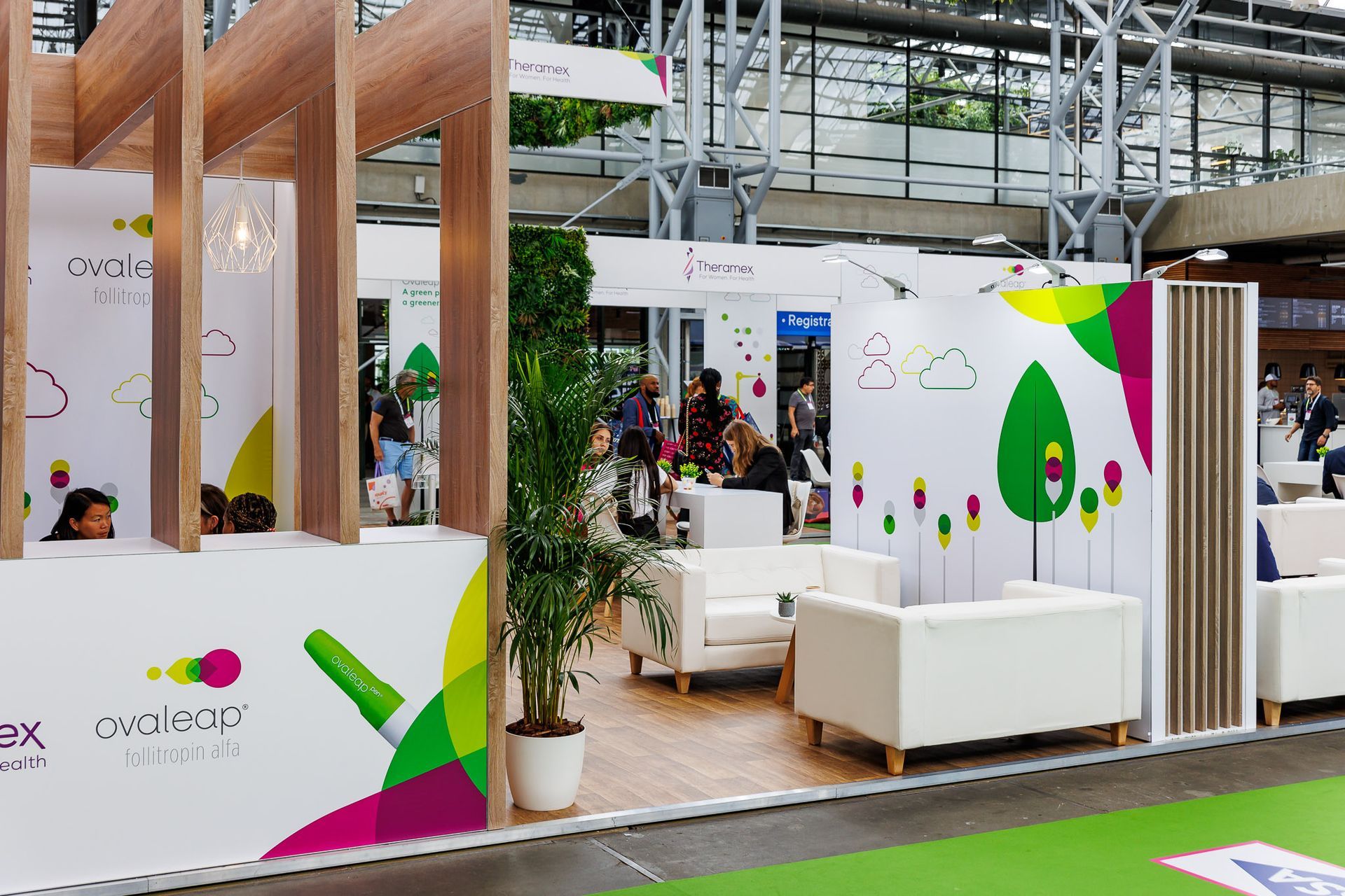 Exhibition booth with white couches, wood accents, and colorful branding. People are visible in the background.