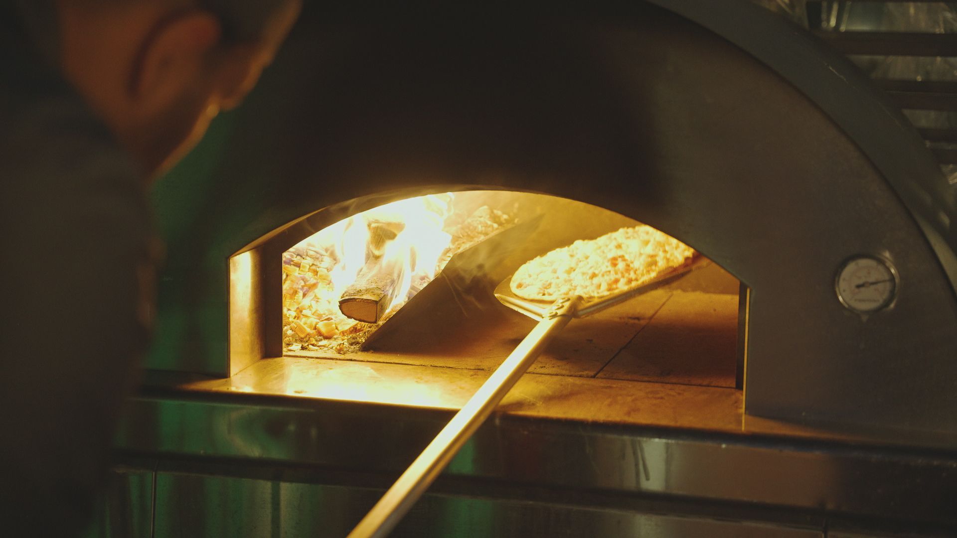 Chef placing pizza in a wood-fired oven. Flames visible, interior setting.