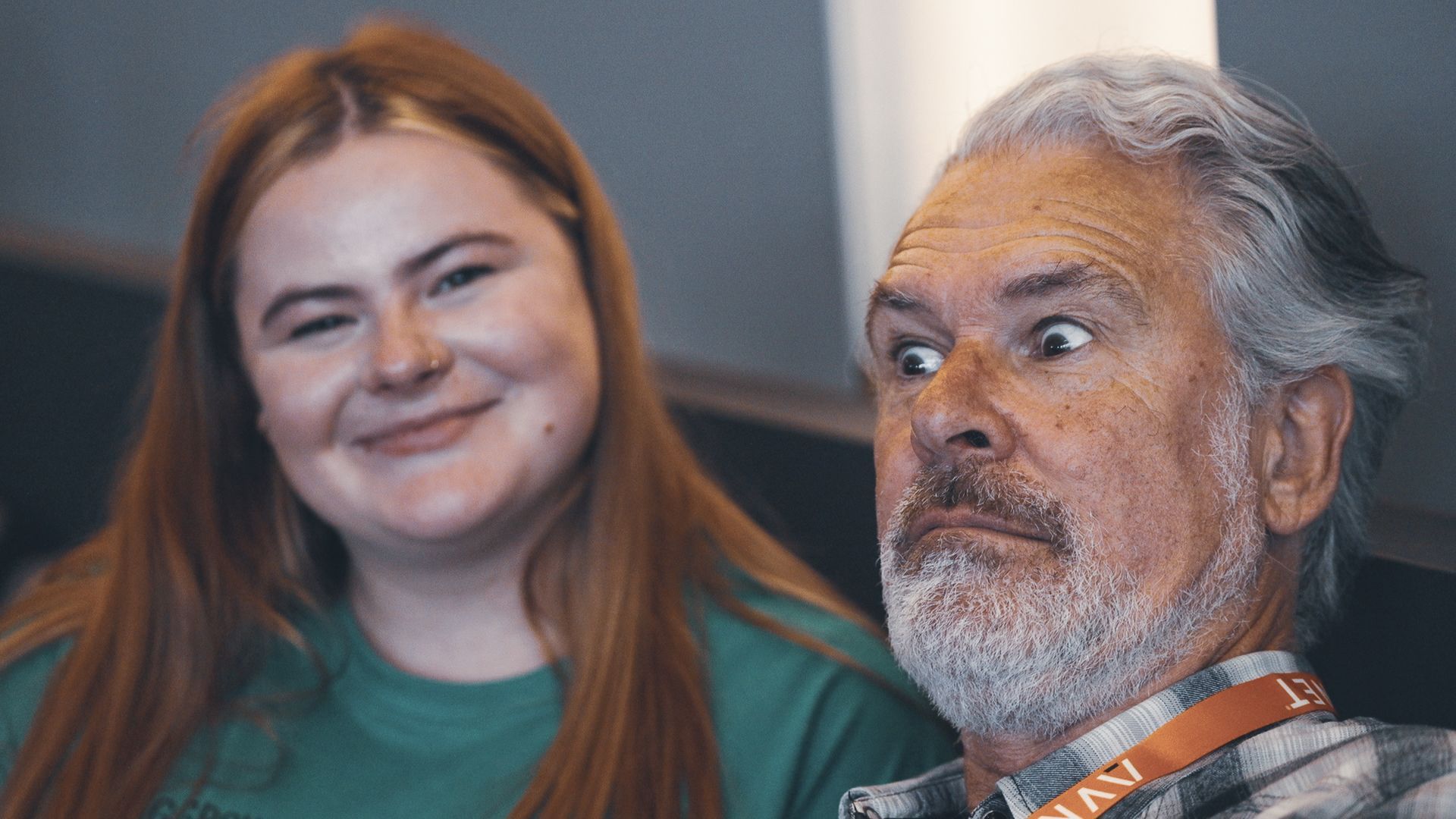 Woman smiles at the camera while an older man makes a shocked expression, indoors.