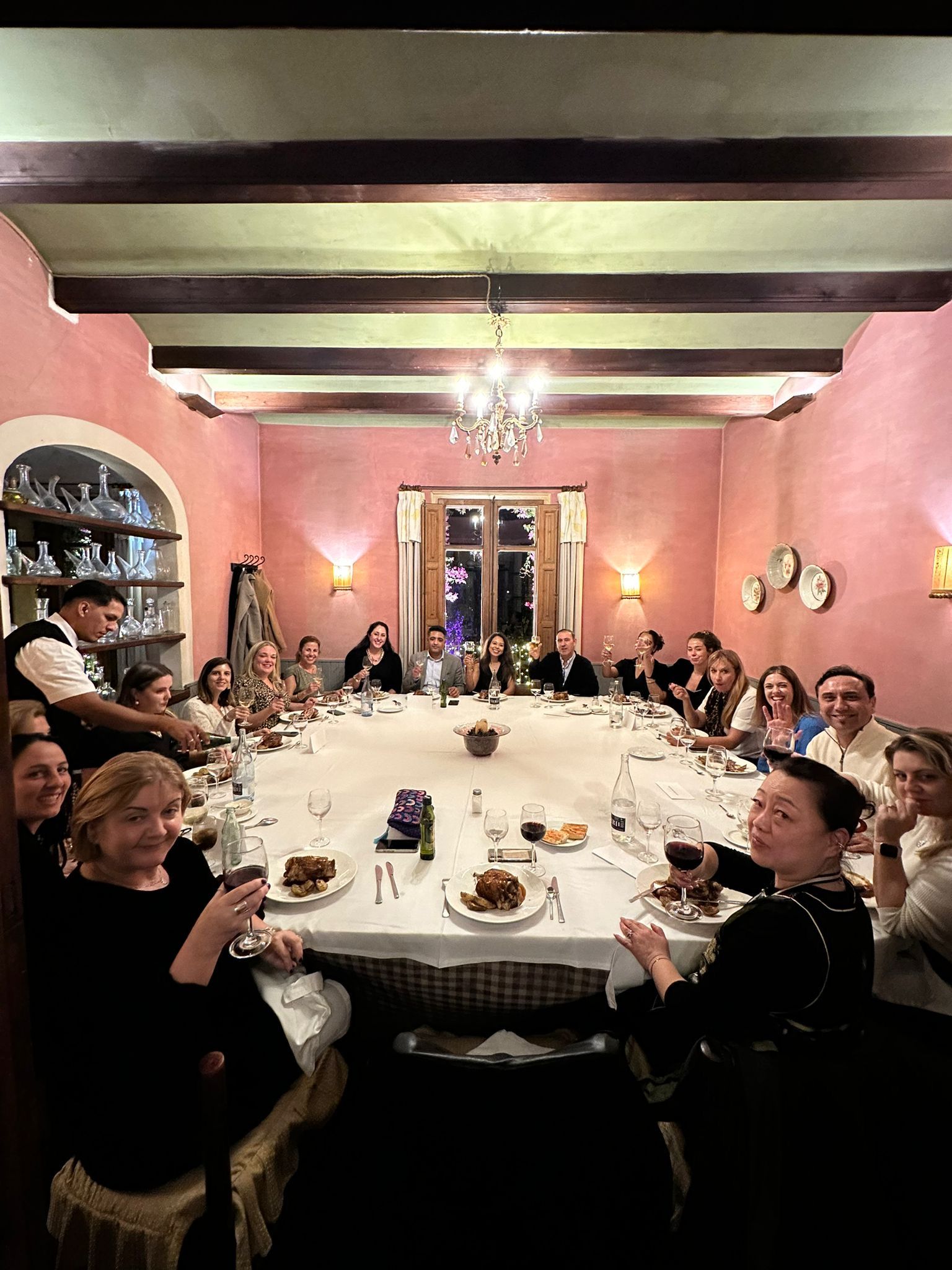 People around long table, raising wine glasses. Pink walls, ornate chandelier, festive setting.