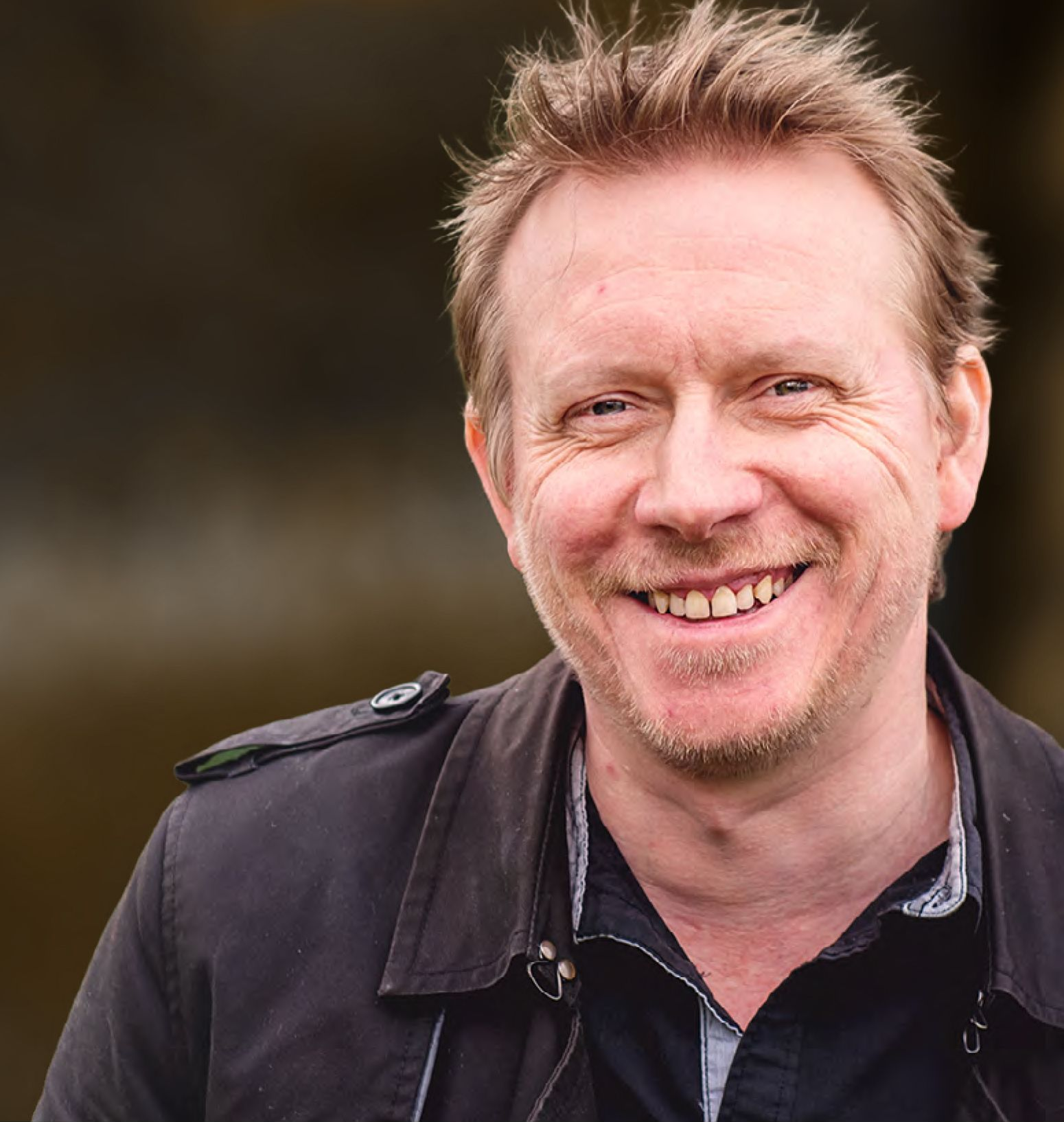Smiling man with blond hair in black jacket outdoors.