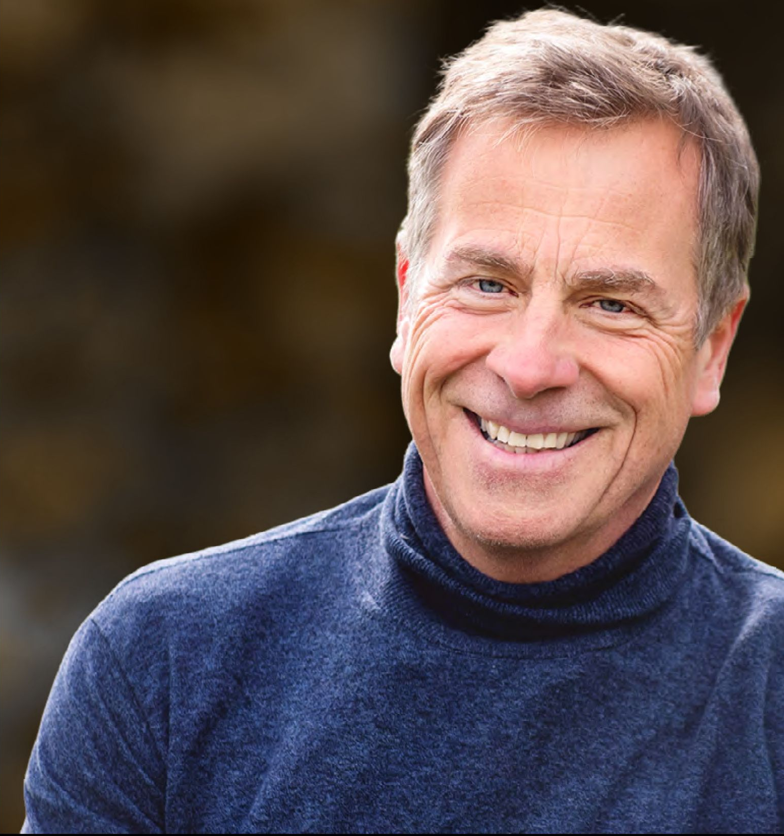 Smiling man with short gray hair in a blue turtleneck; outdoors with greenery and a dirt path in the background.