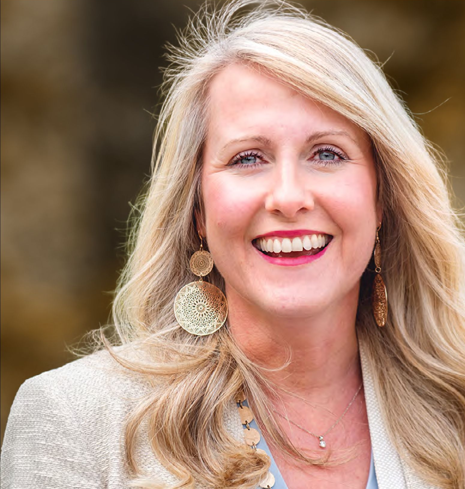 Smiling woman with blonde hair wearing gold earrings and a blazer outdoors.