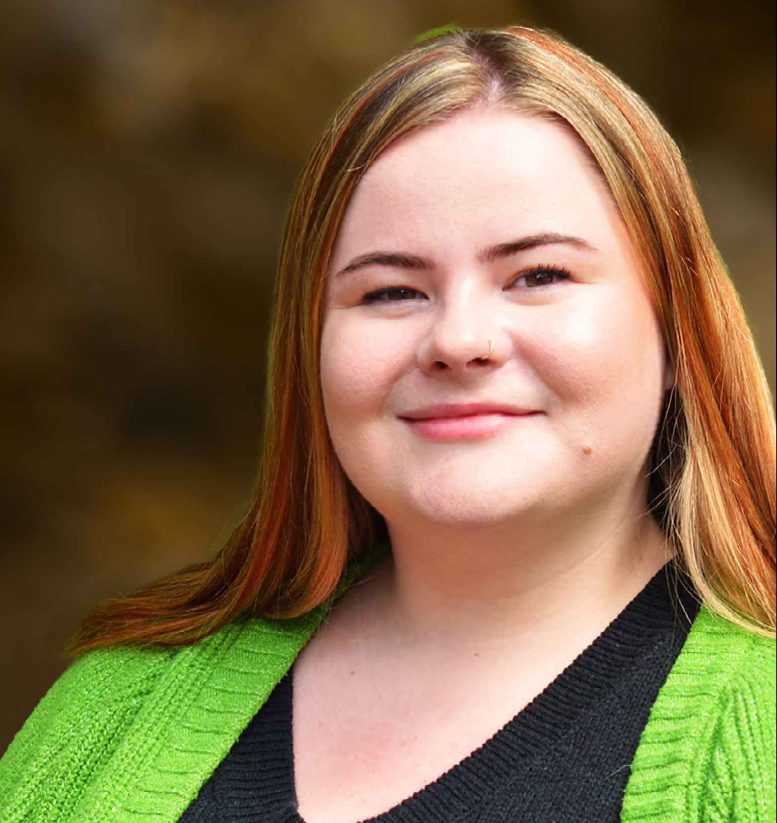 Woman with long red-blonde hair smiling, wearing a green cardigan and black top, outdoors in front of greenery.