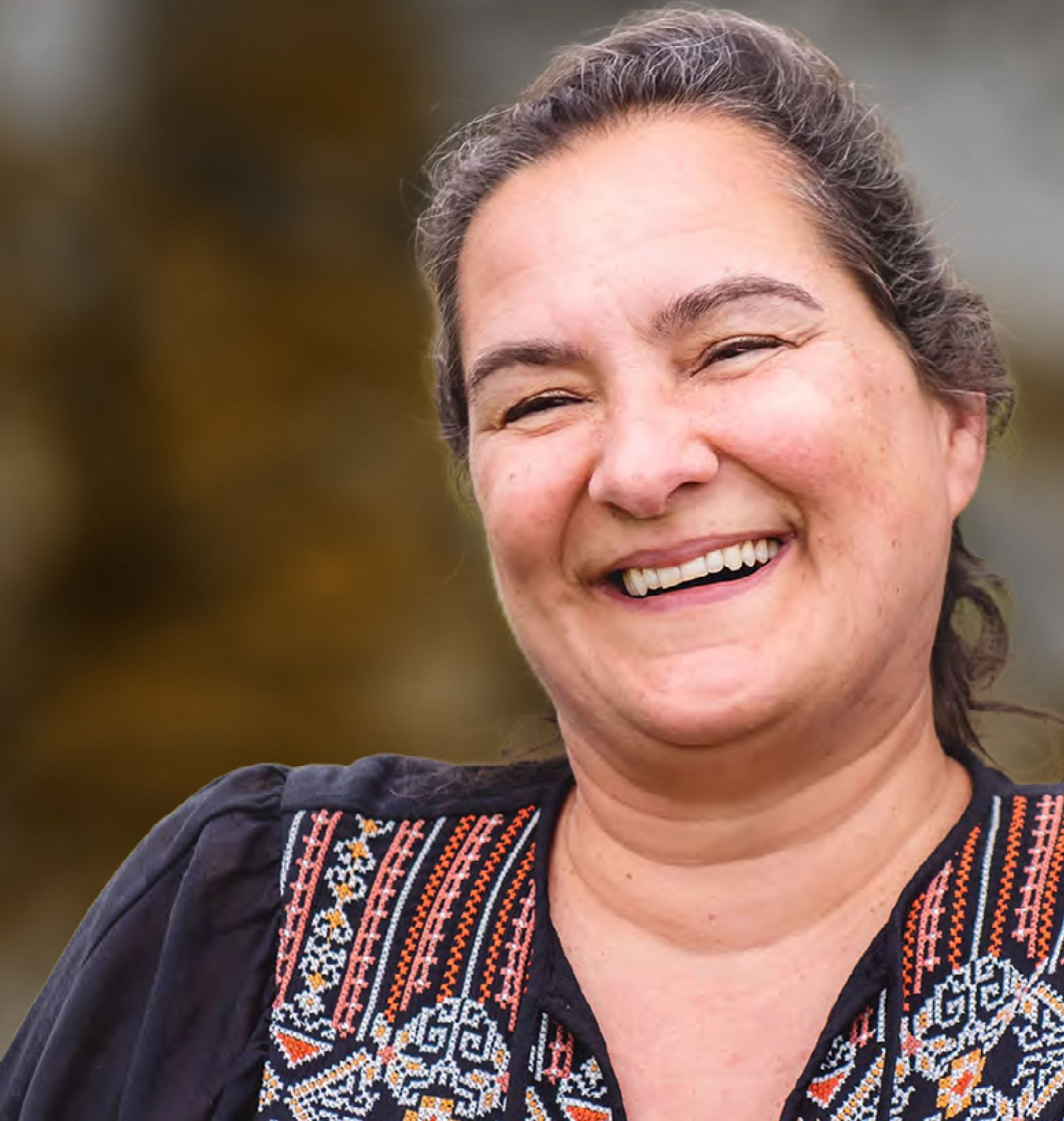 Woman with dark hair and embroidered shirt smiling outside.