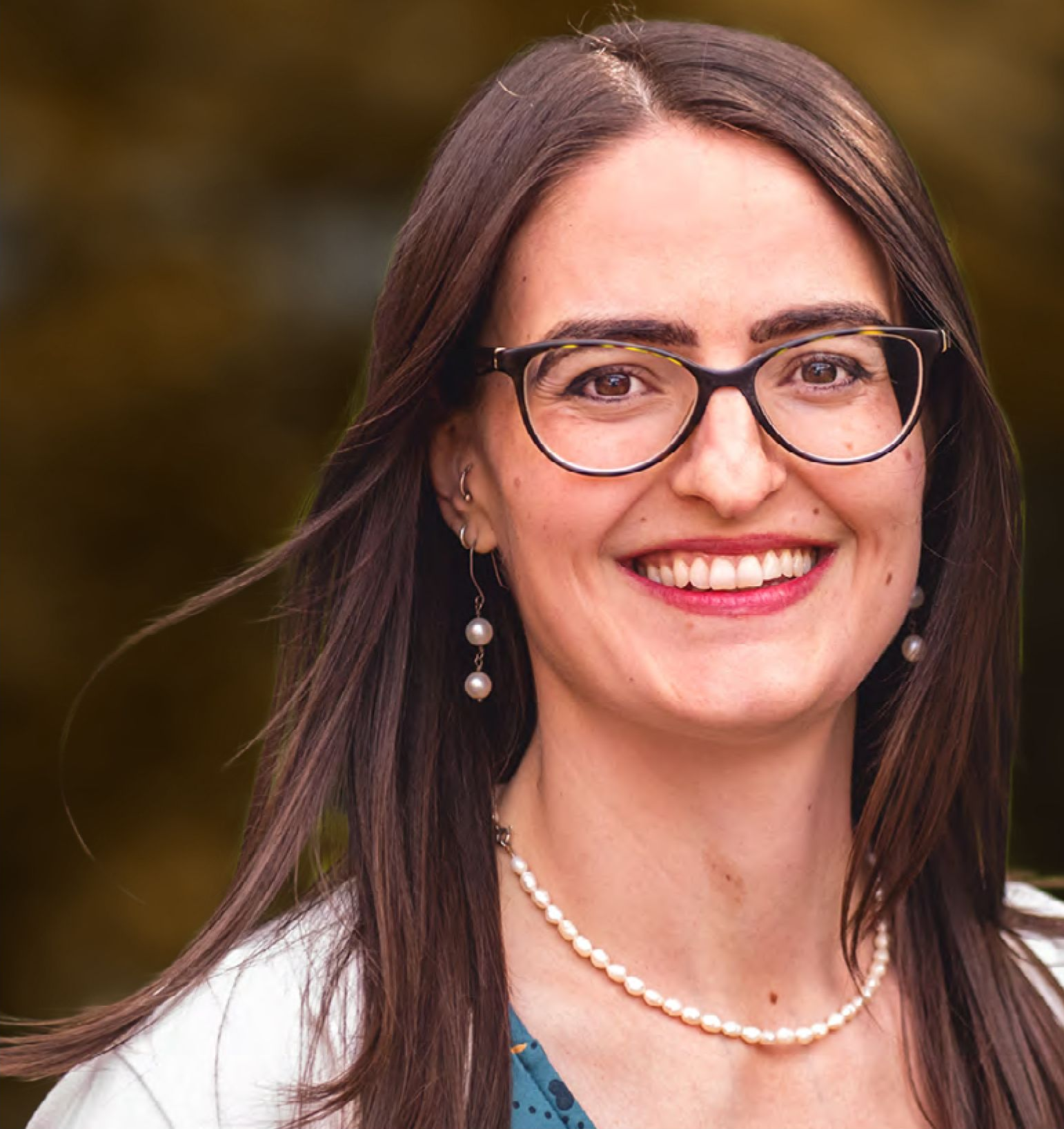 Woman with glasses, smiling, wearing pearl necklace and earrings, in front of green foliage.