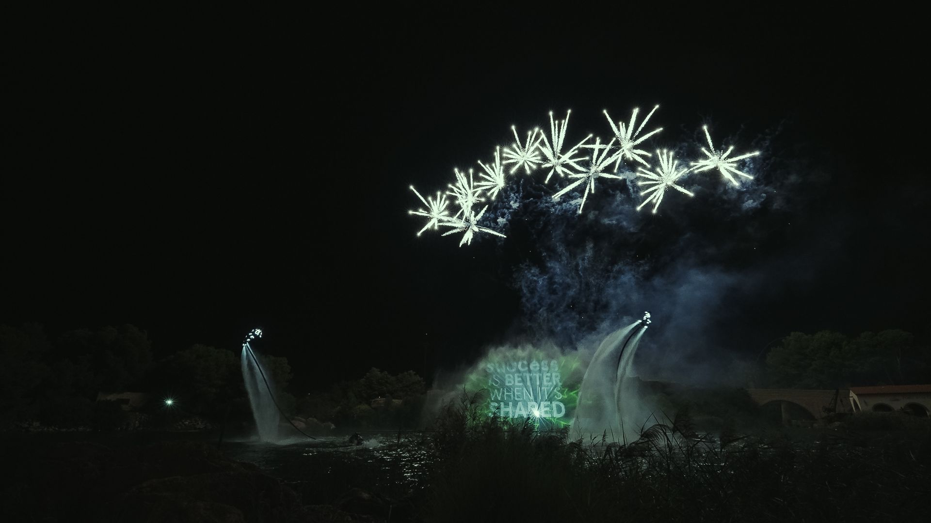 Fireworks burst over a fountain display at night, with white lights against a dark sky.