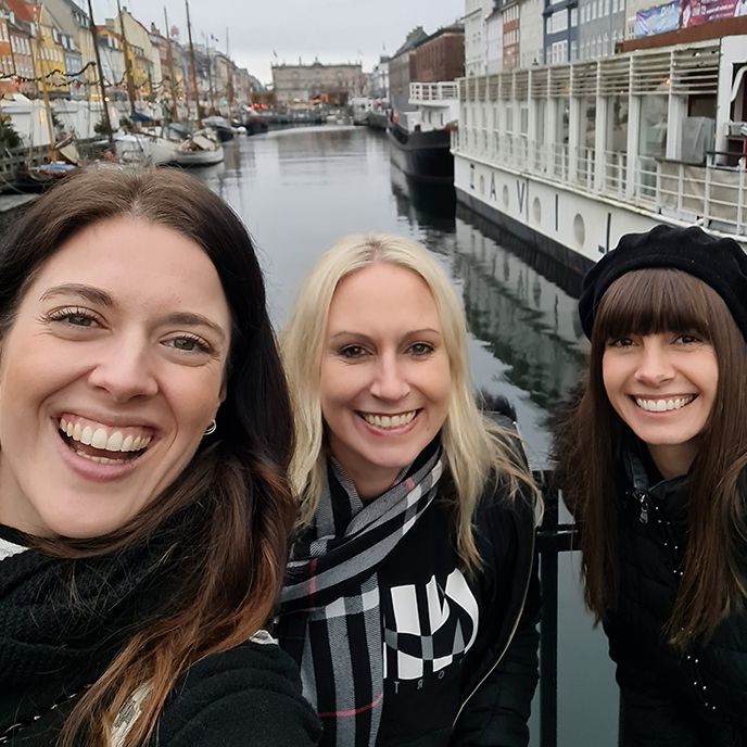 Three smiling women take a selfie on a bridge in Copenhagen. Buildings and boats line the canal.