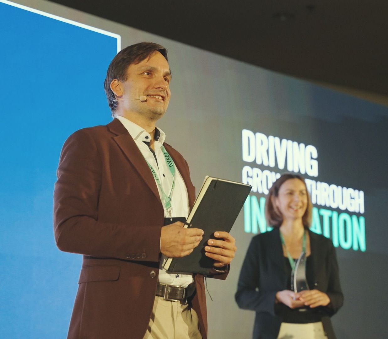 Man in a burgundy jacket holding a tablet smiles on stage with a woman. Text: 