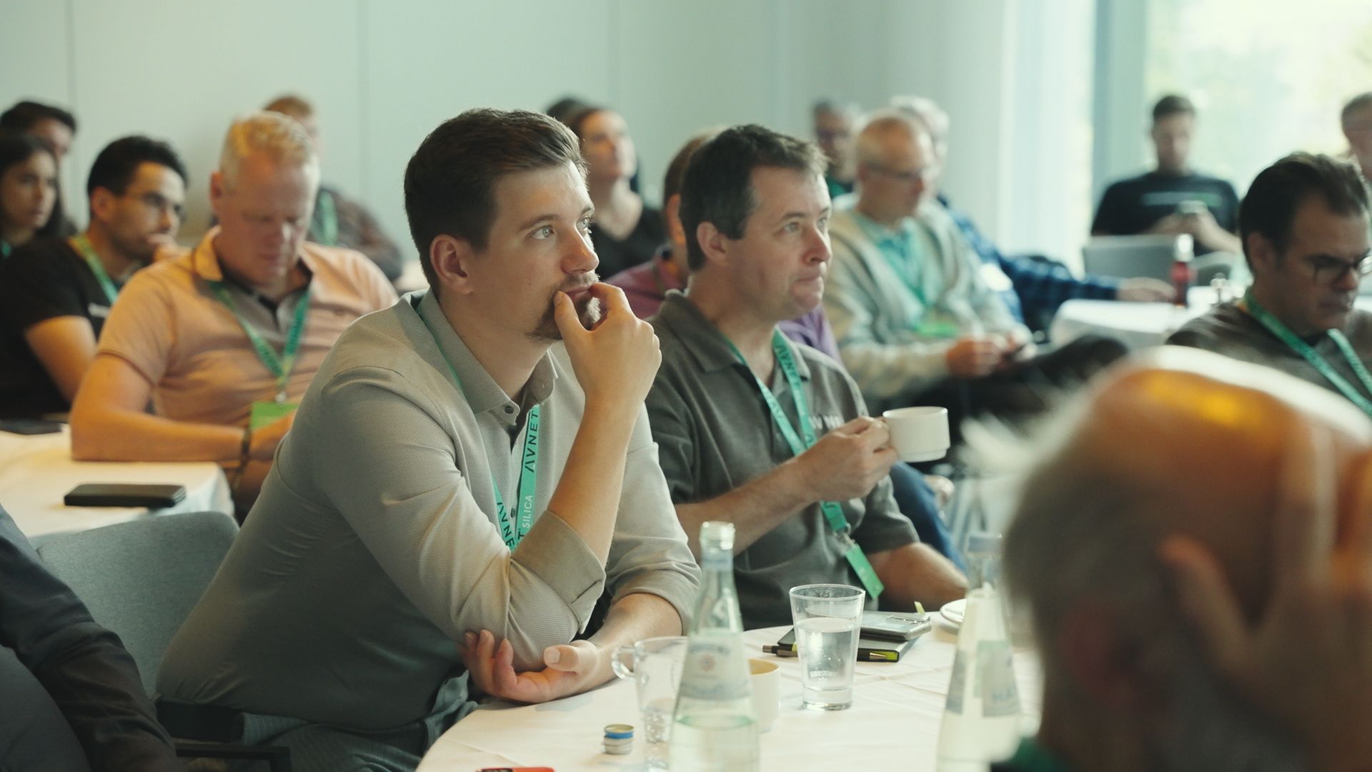 Audience at a conference, focused on a speaker. People are diverse, mostly middle-aged, wearing lanyards, indoors.