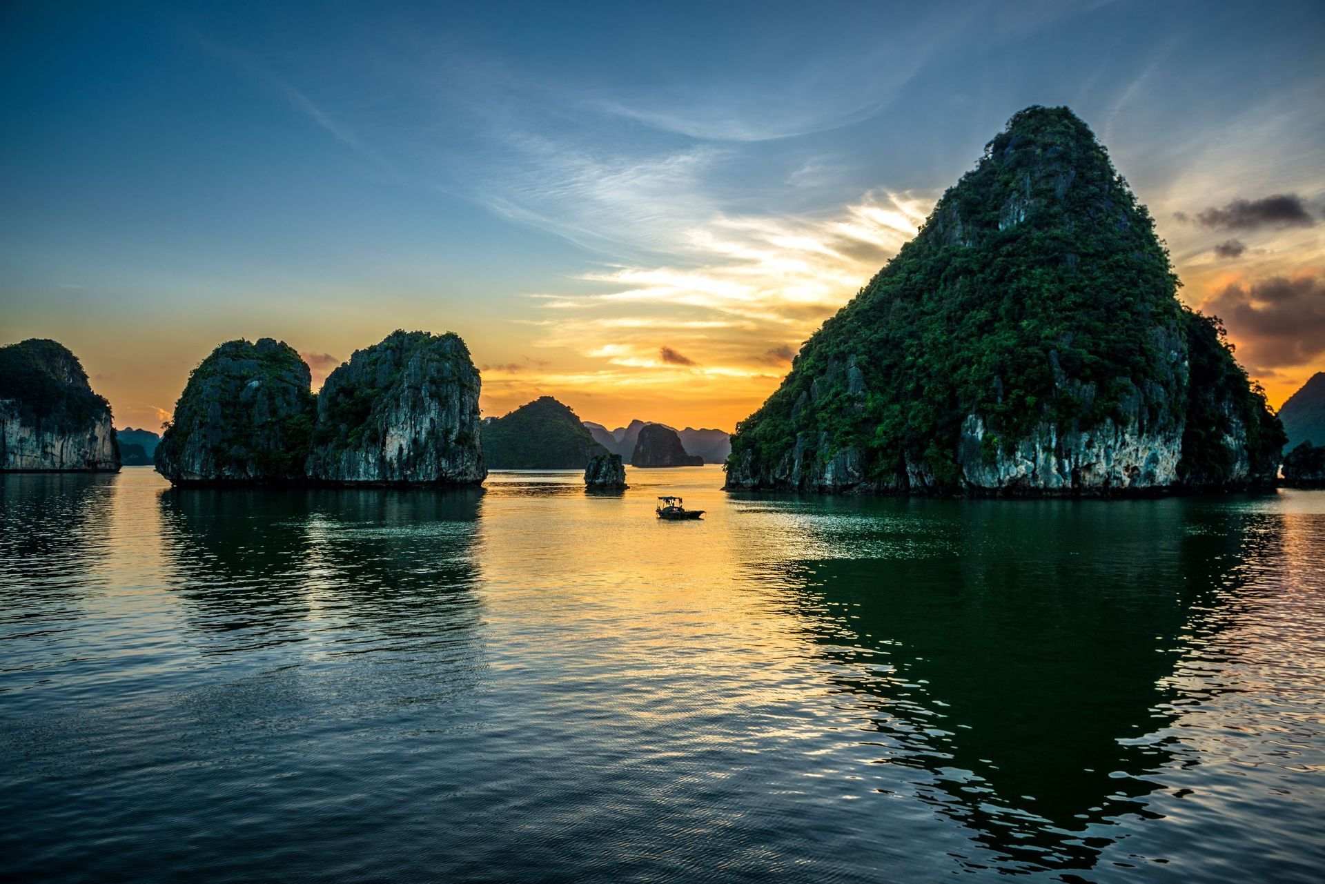 Sunset over Ha Long Bay, Vietnam; limestone karsts rise from calm water, small boat in the distance.