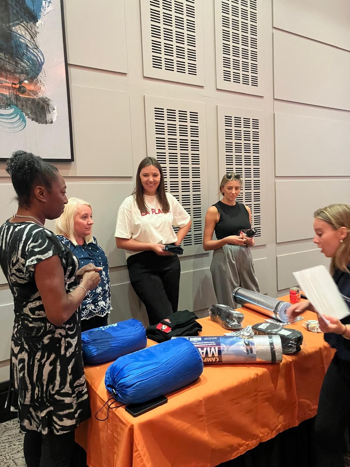 Five people at a table with camping gear. An orange table cloth, white wall background.