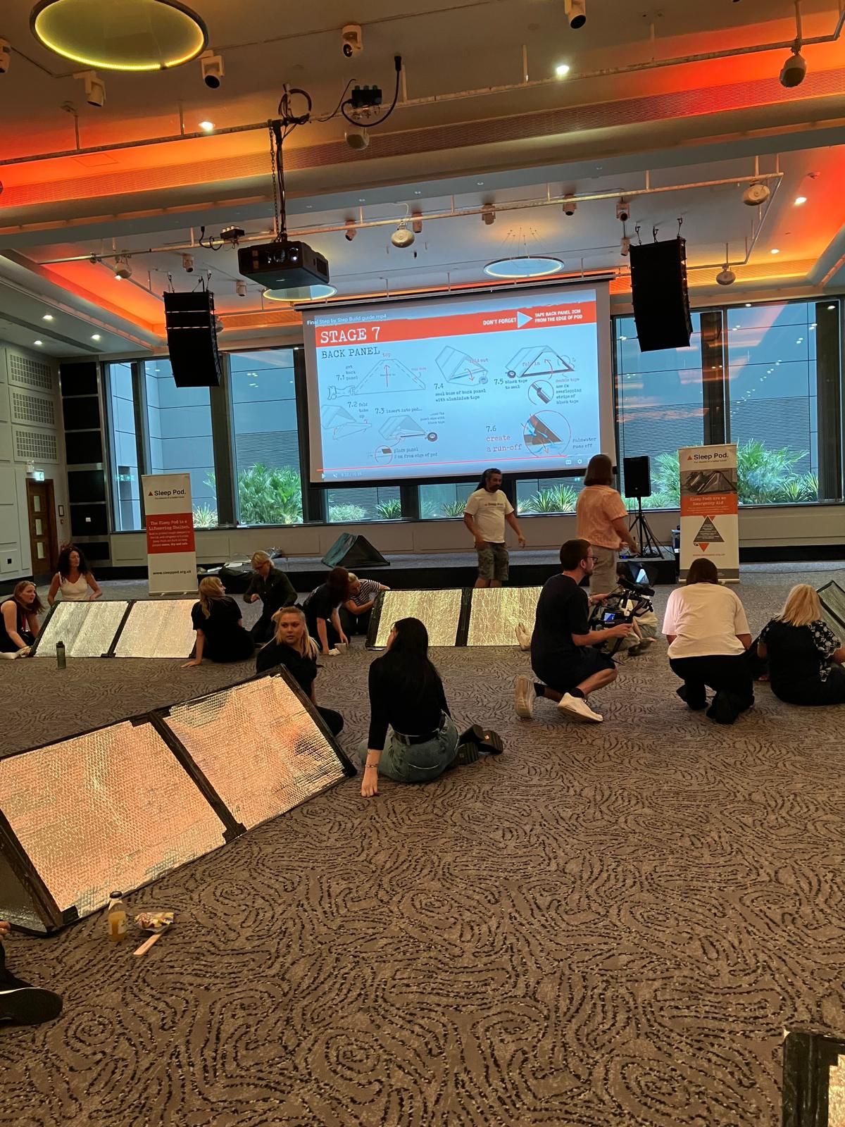 People in a conference room, looking at a presentation on a screen. Some are on the floor, looking at panels.