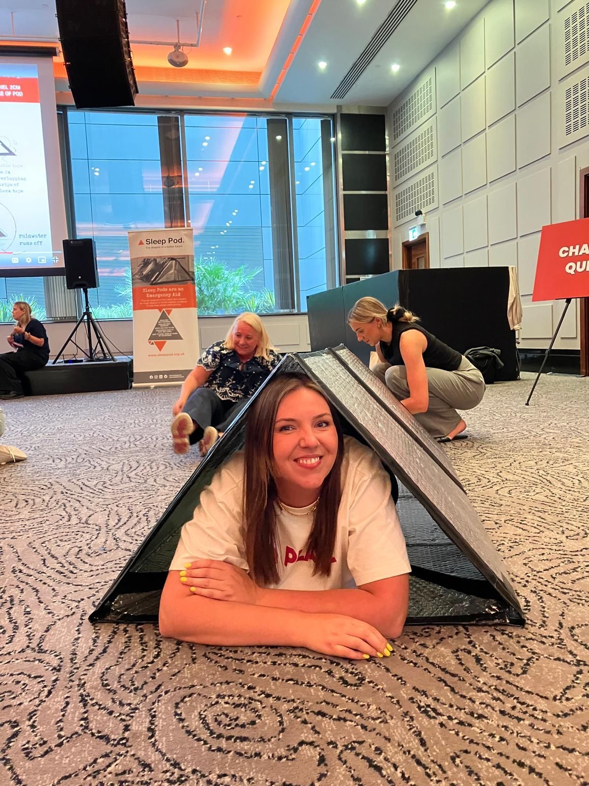 Woman smiles, peering out from a small, black tent-like structure, with two others crouching behind her in a meeting room.