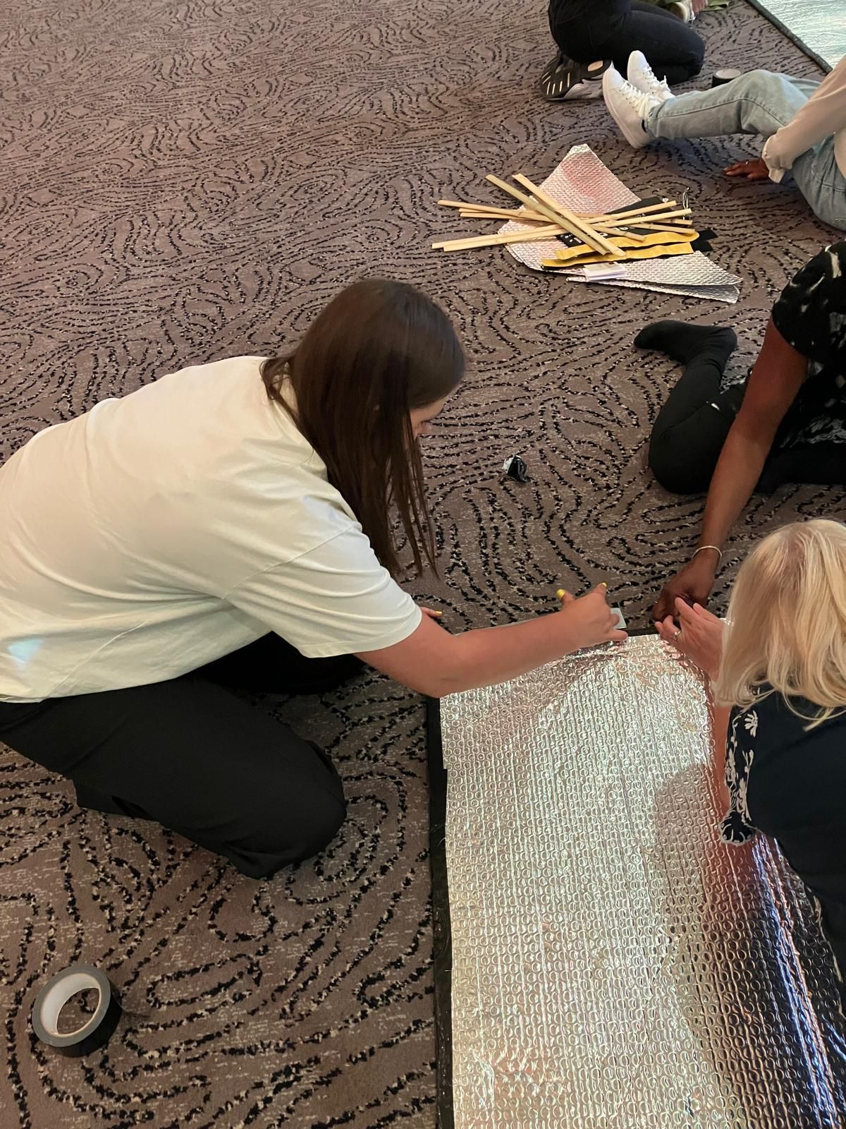 People kneeling on carpet, assembling reflective material with tape.