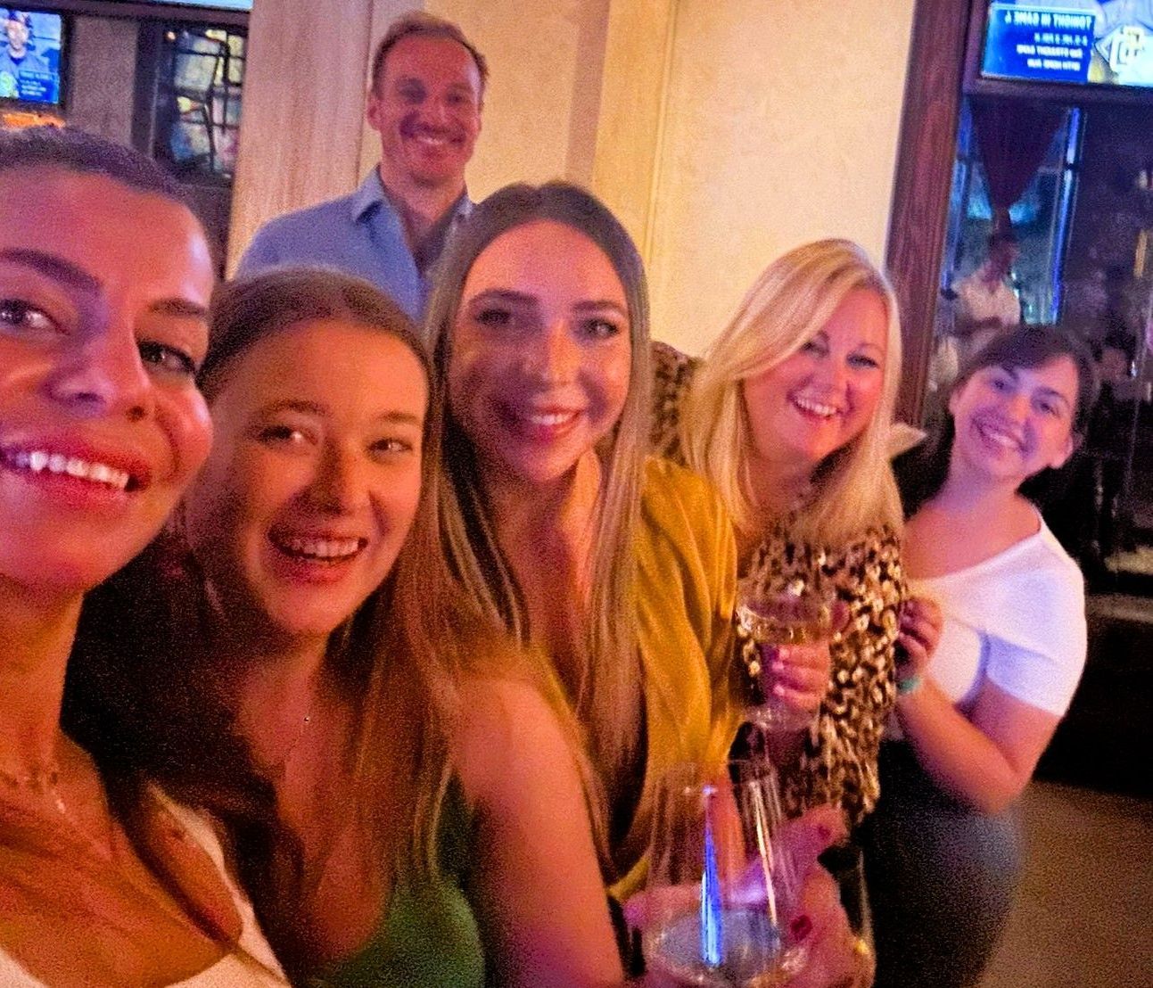 Group of smiling people at a bar, posing for a photo. Some holding drinks, joyful expressions.