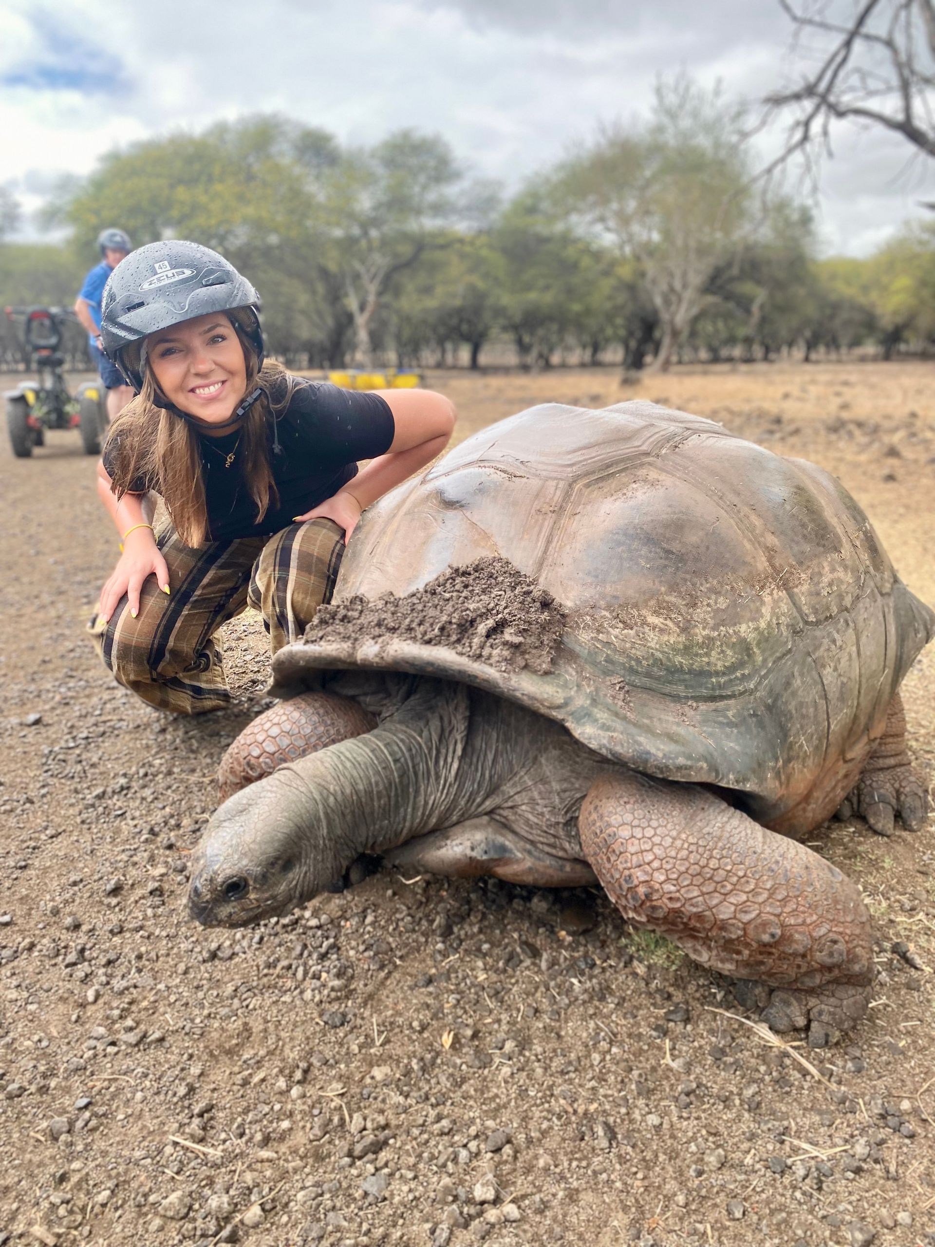 Woman smiles next to a giant Galapagos tortoise. They are outdoors on a dirt path with a vehicle in the background.
