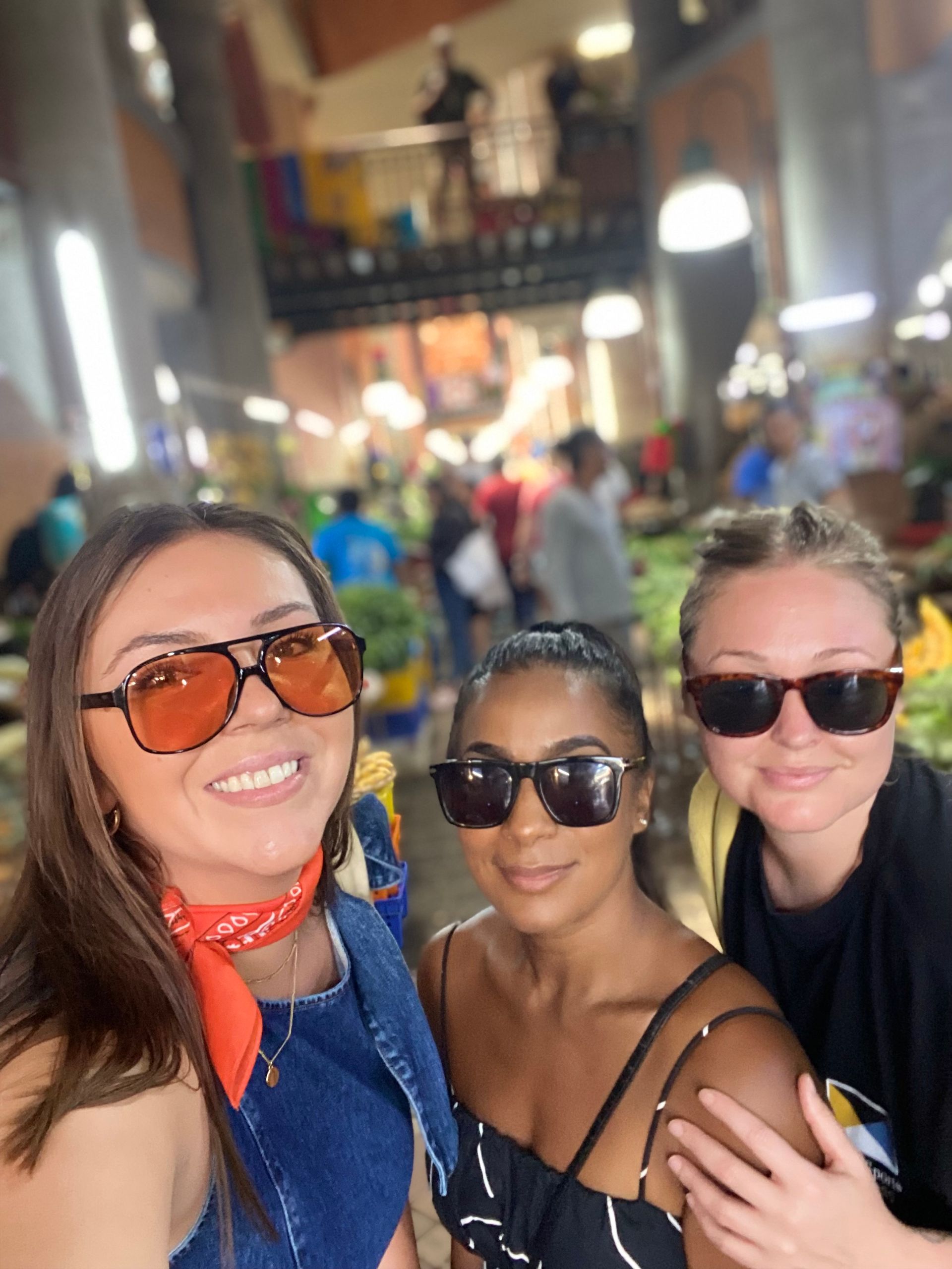 Three women in sunglasses smile at the camera in a bustling market.